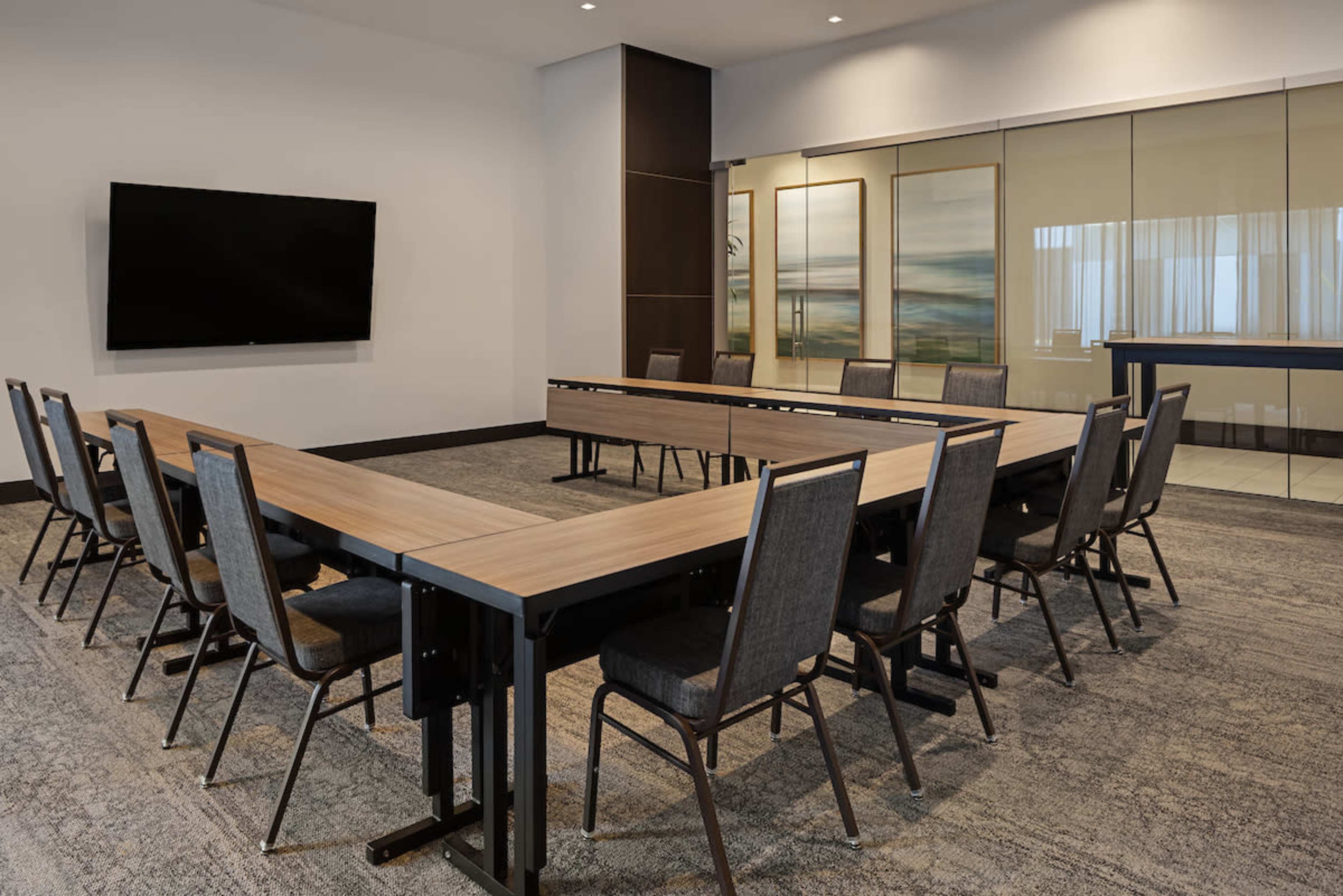 The image shows a conference room set up with tables arranged in a U-shape, surrounded by chairs, and a large screen mounted on the wall.