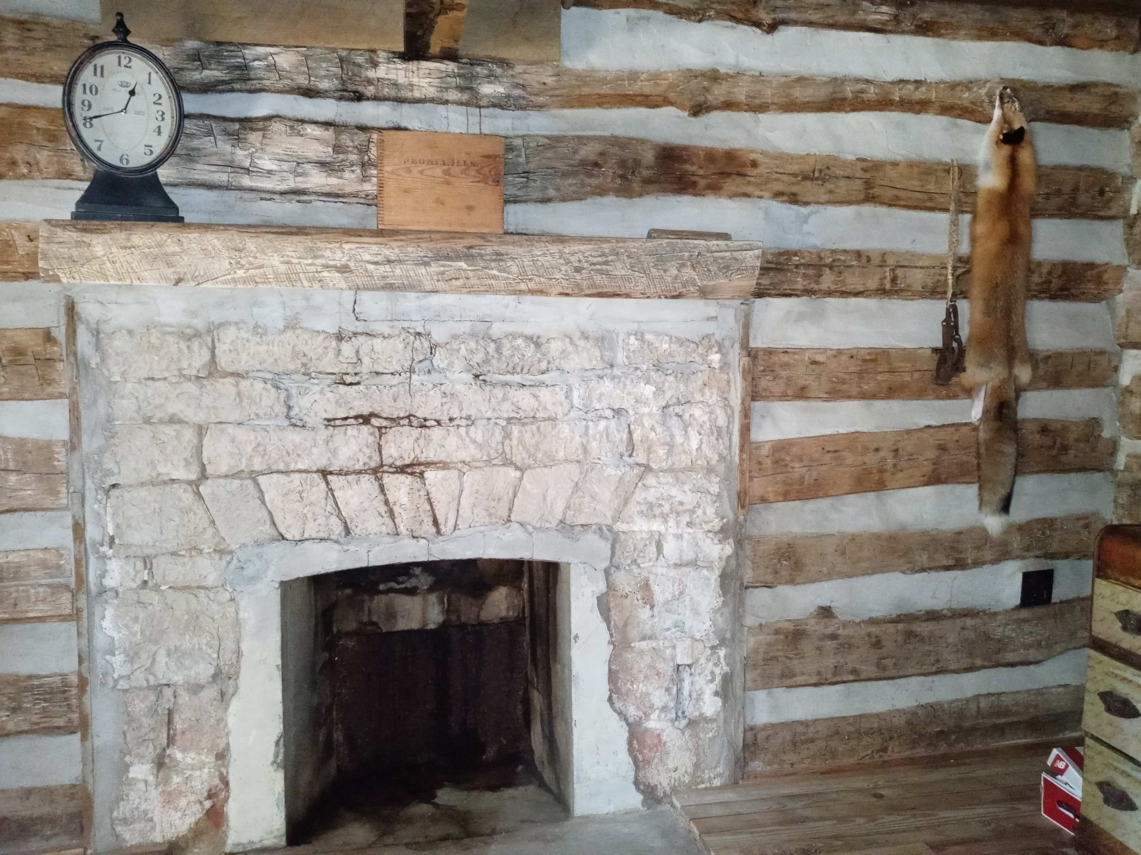 The image shows a rustic room with a stone fireplace, wooden logs forming the walls, and a clock mounted on the wall.