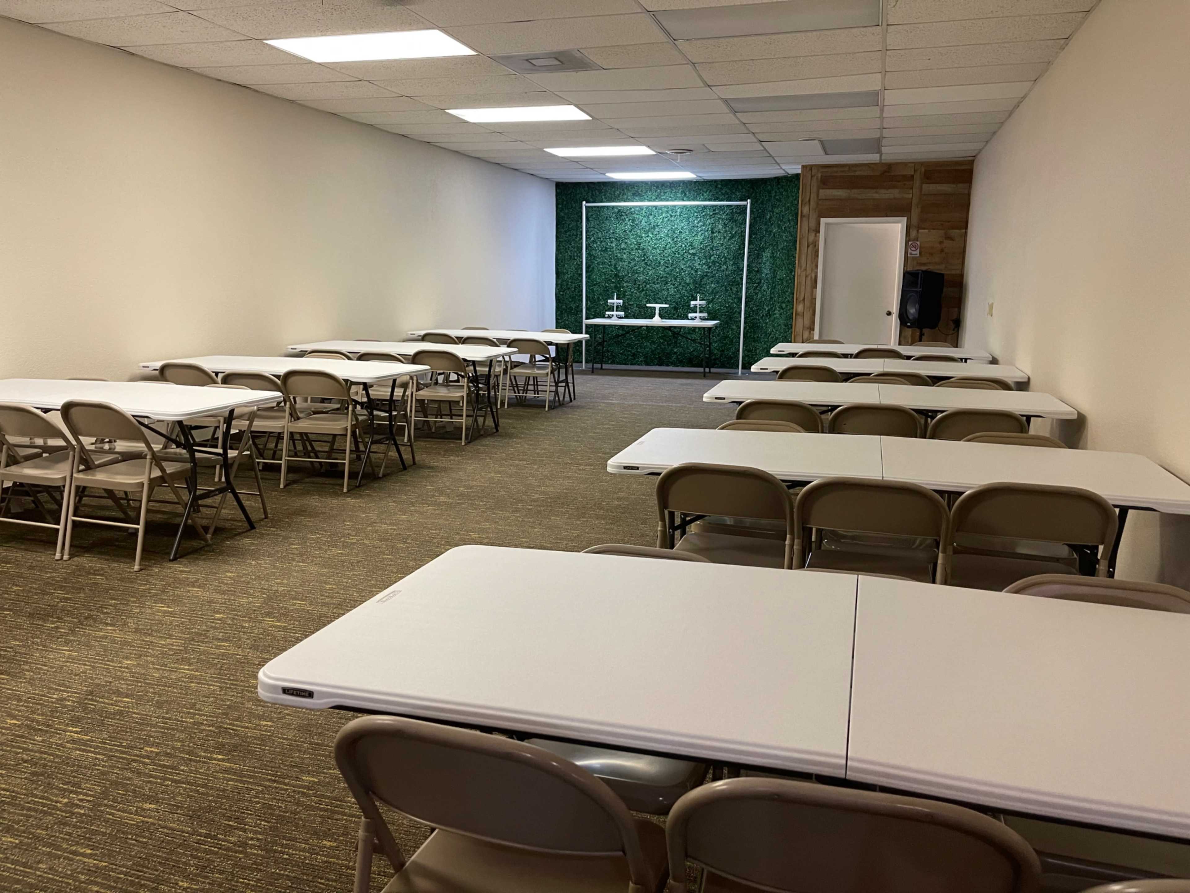 An empty meeting room with multiple folding tables and chairs arranged in rows, a green wall feature in the background, and a door on the right side.