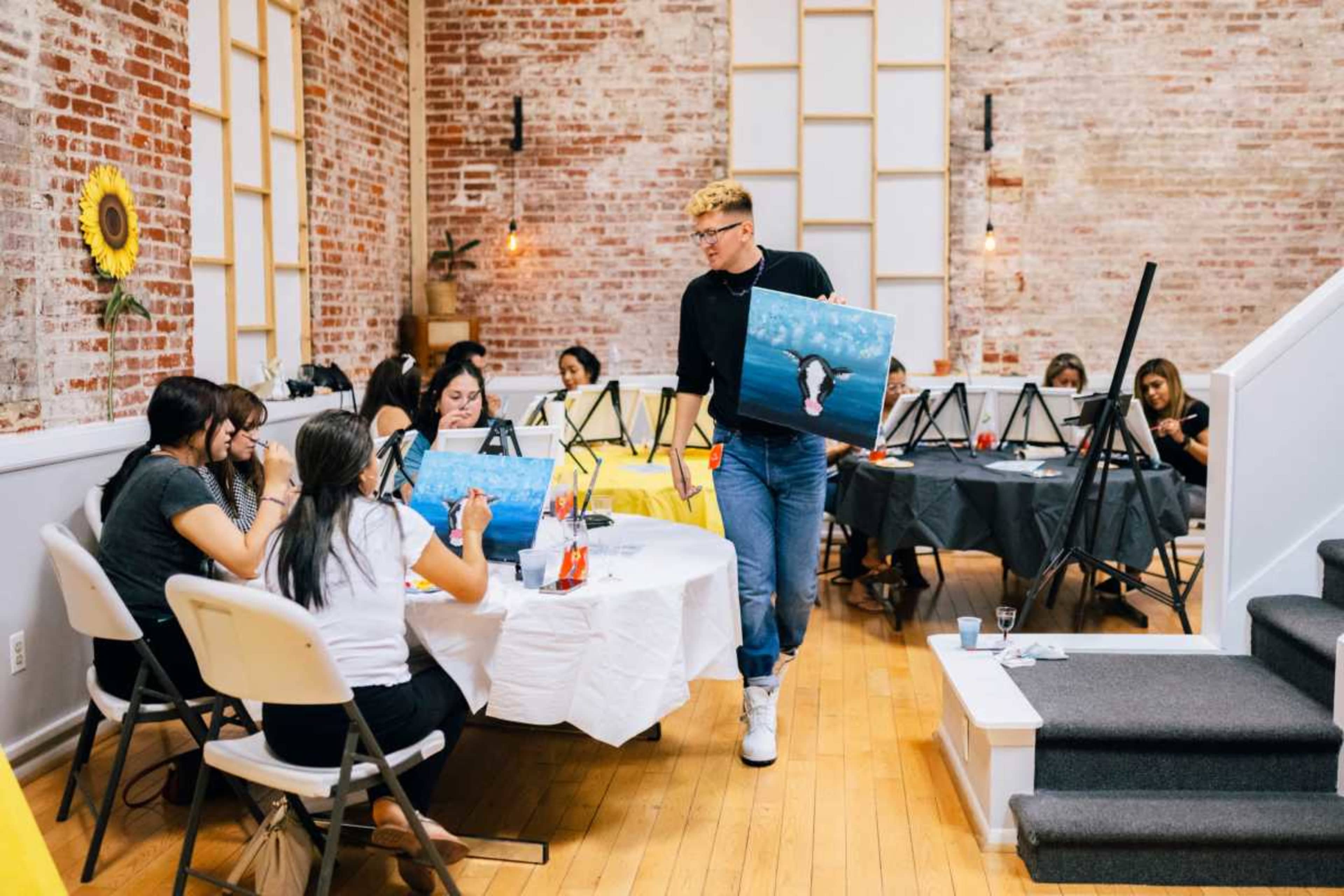 A group of people are painting on canvases during a class in a brightly lit studio with exposed brick walls.