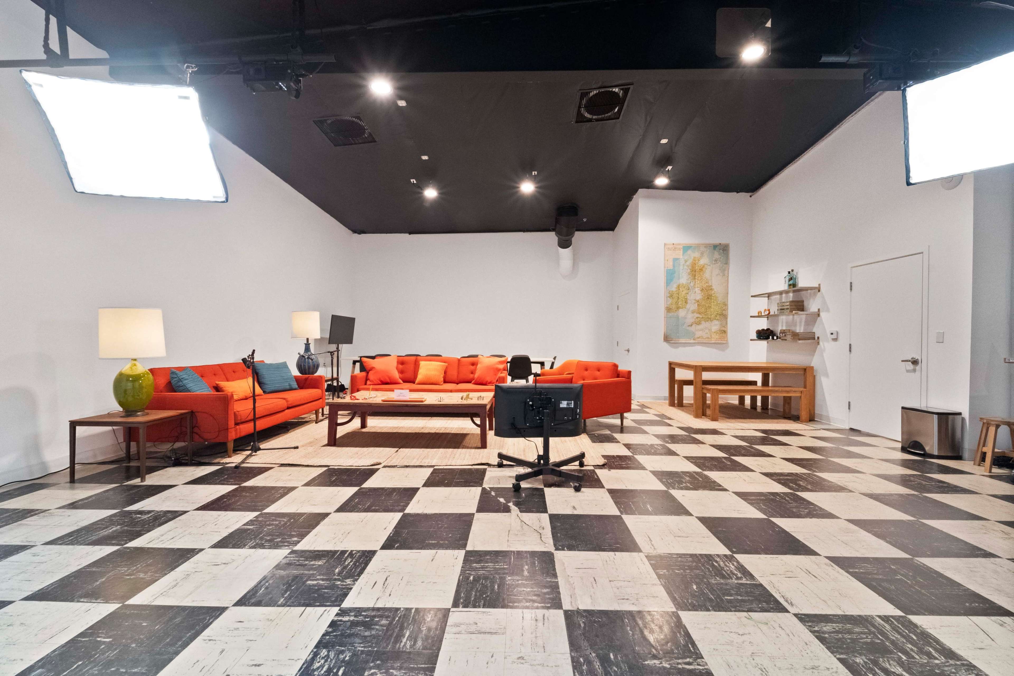 A brightly lit studio space featuring a large orange sectional sofa, a wooden coffee table, and a checkered black and white floor.