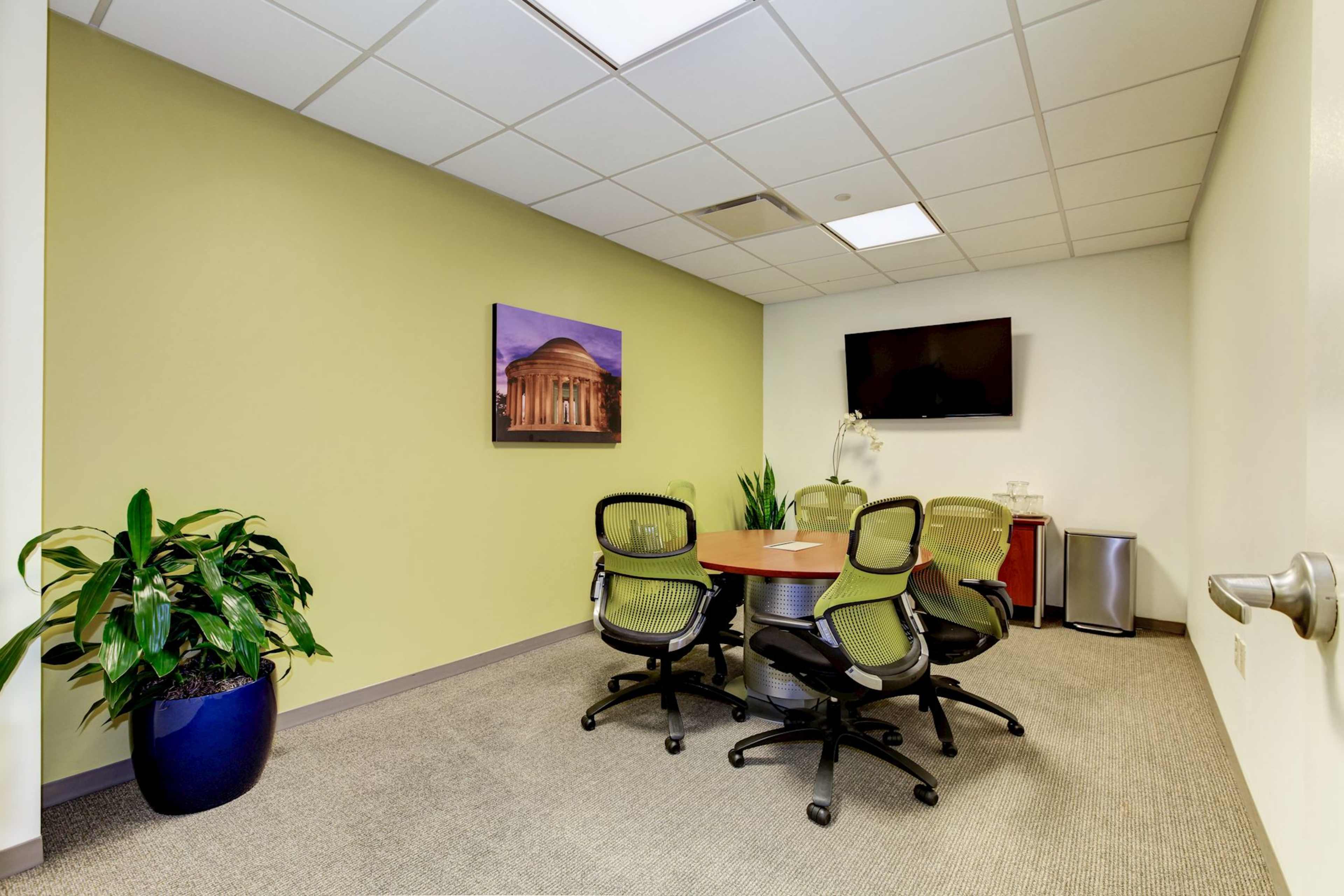 A small meeting room with a round table surrounded by four chairs, a green plant in a blue pot, a flat-screen TV on the wall, and a framed picture of a building.