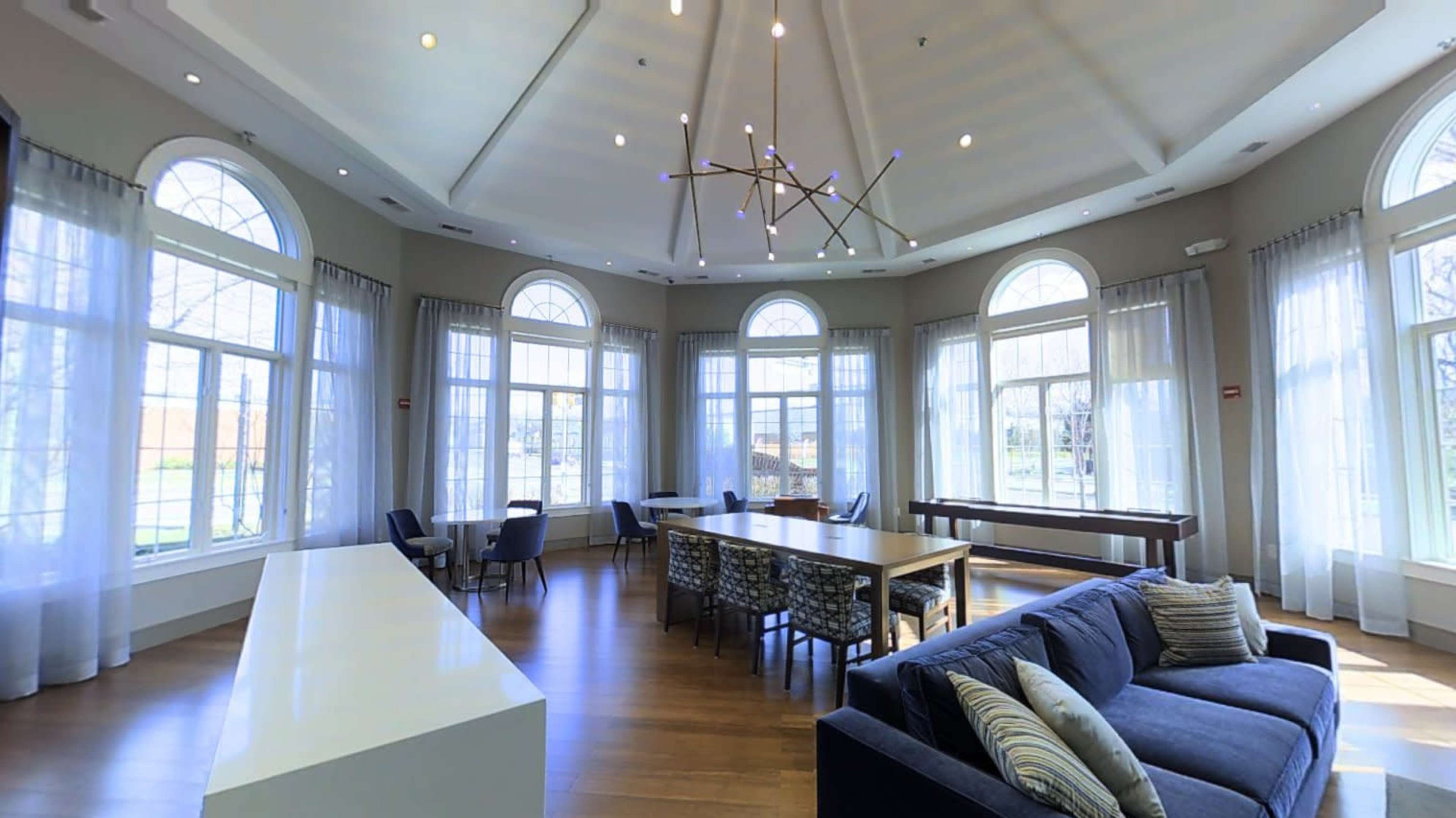 A spacious room with large windows, a seating area with a sofa, and a dining table under a decorative ceiling fixture.