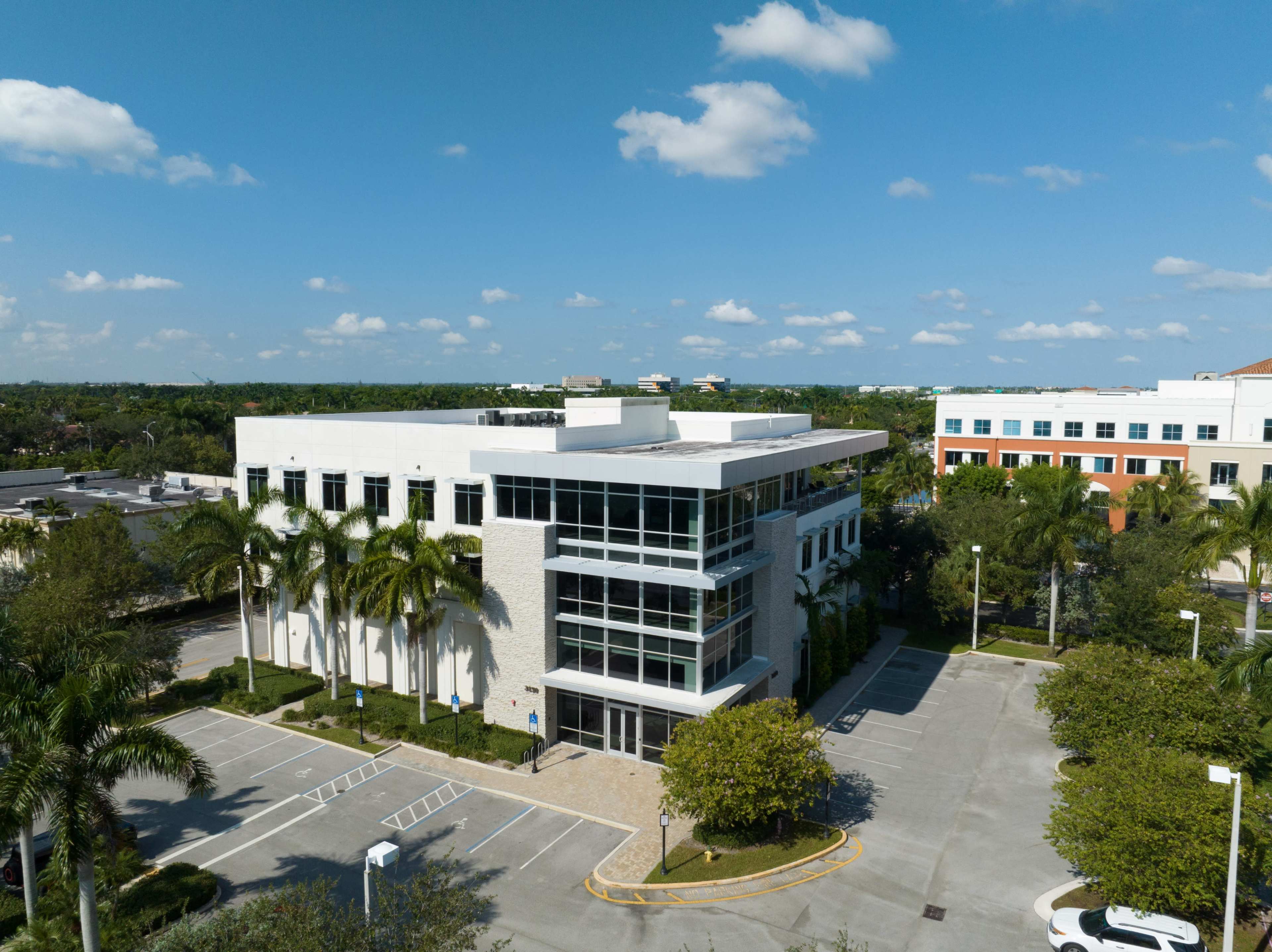 A modern office building with large windows is surrounded by palm trees and an empty parking lot under a clear blue sky.
