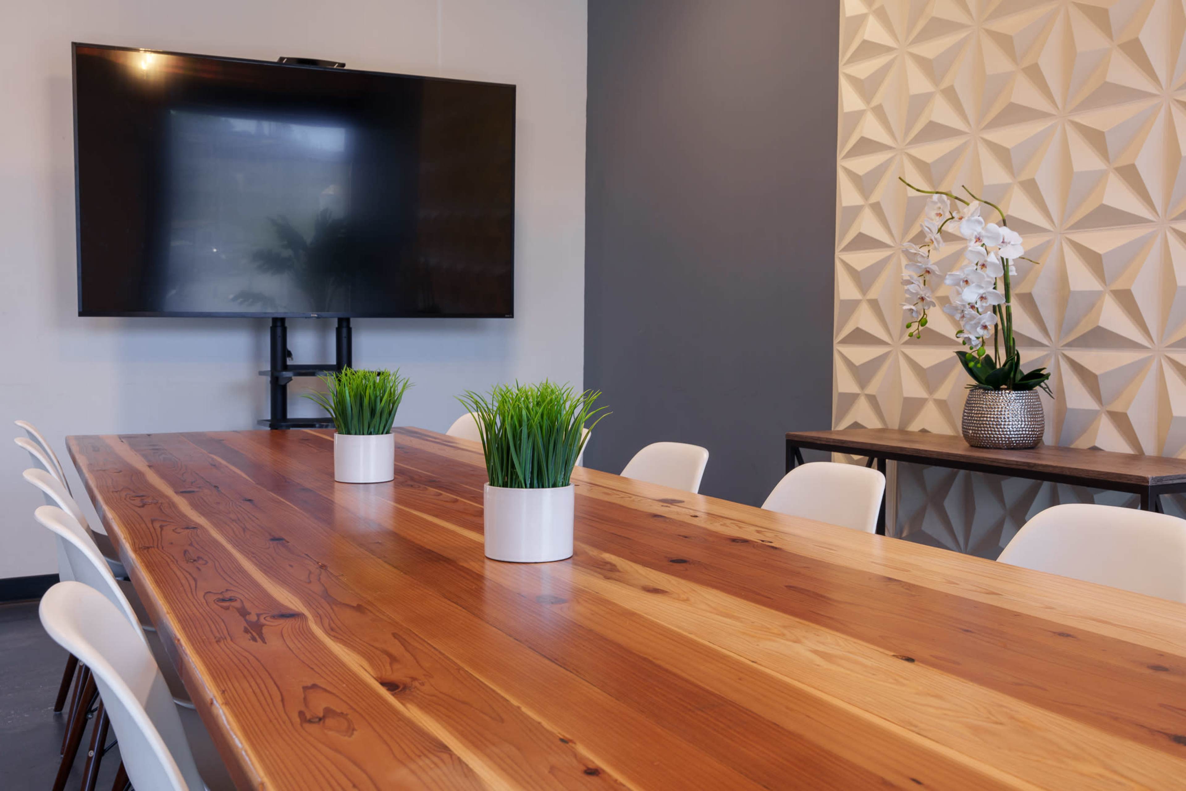 A large wooden conference table with white chairs is set in a meeting room featuring a wall-mounted television and decorative plants.