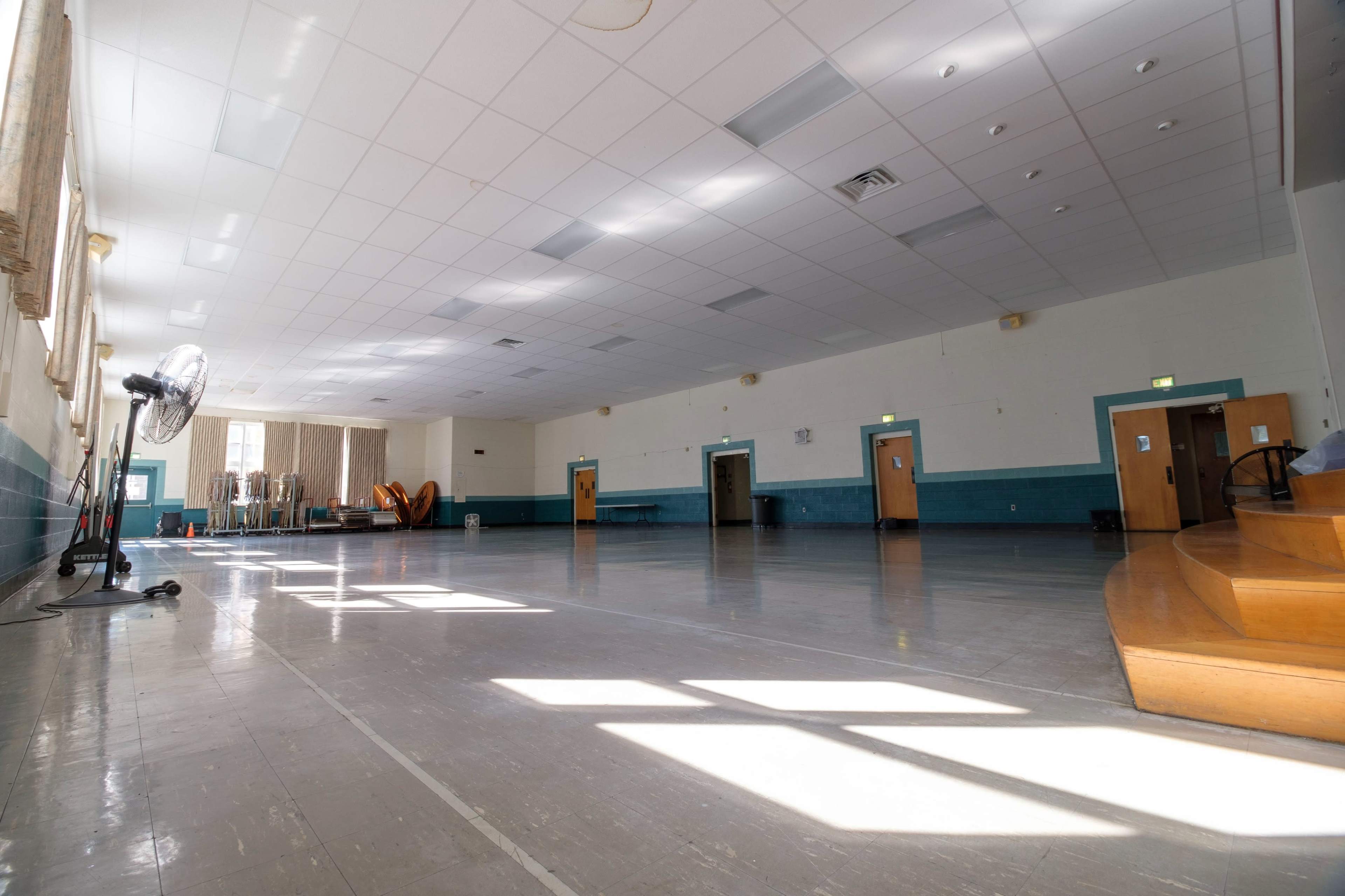 A large, empty indoor space with a shiny floor, multiple doors, and natural light streaming through the windows.