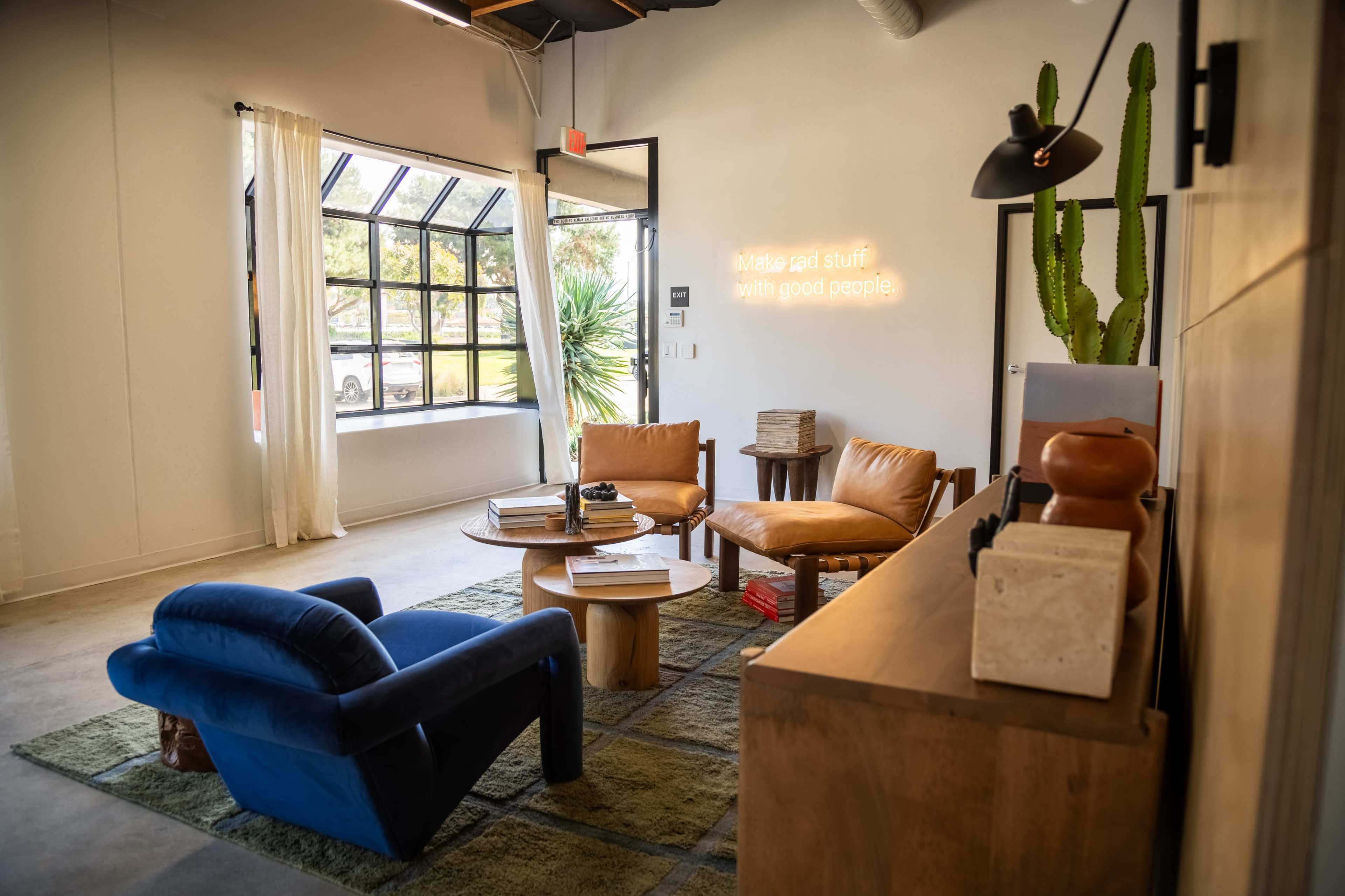 A modern, minimalist lounge area featuring a blue armchair and brown leather chairs arranged around a wooden coffee table, with a cactus and large windows in the background.