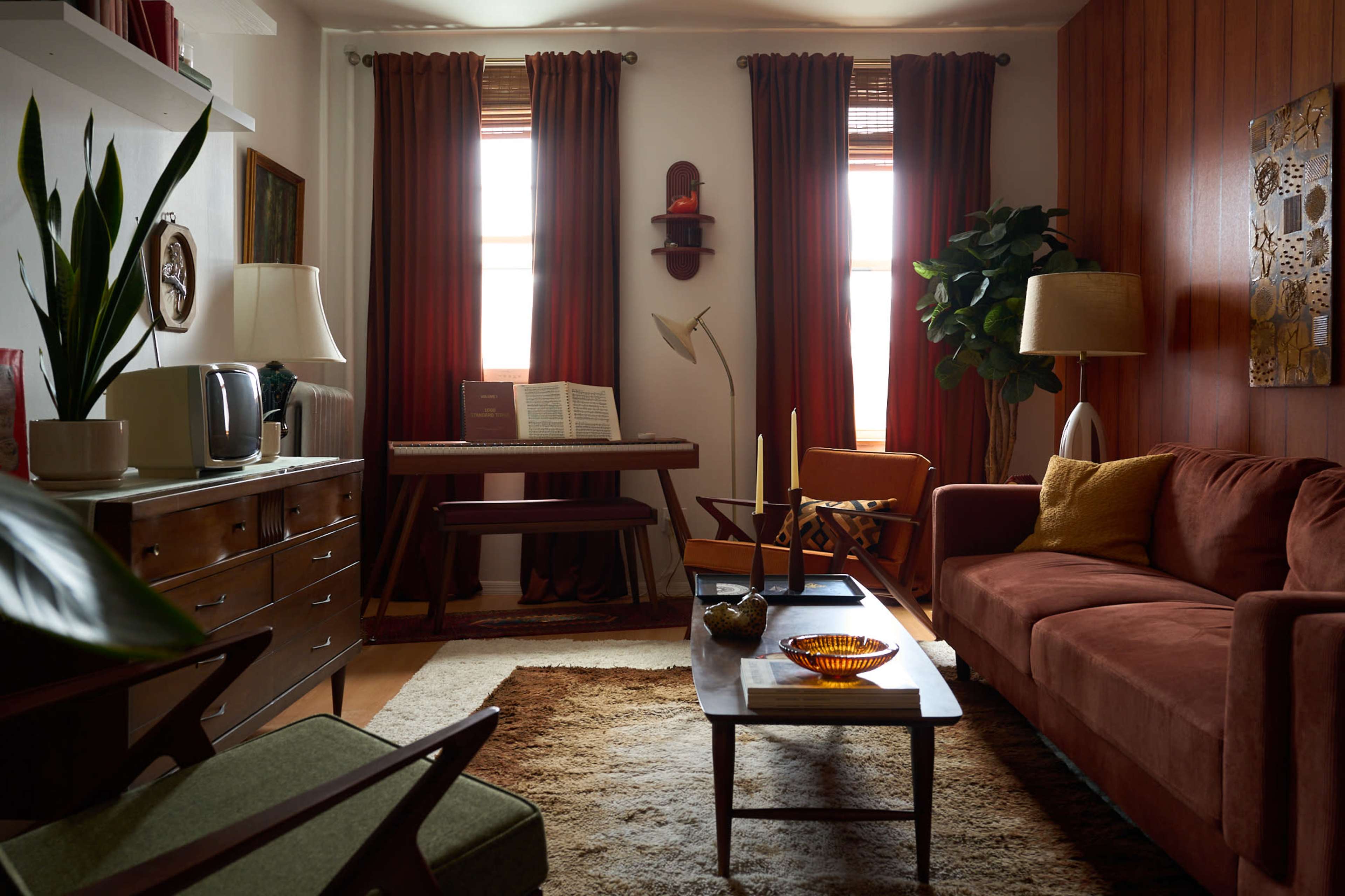 The image shows a cozy living room featuring a brown sofa, a wooden coffee table, a keyboard on a stand, and various plants beside soft natural light coming through the curtains.