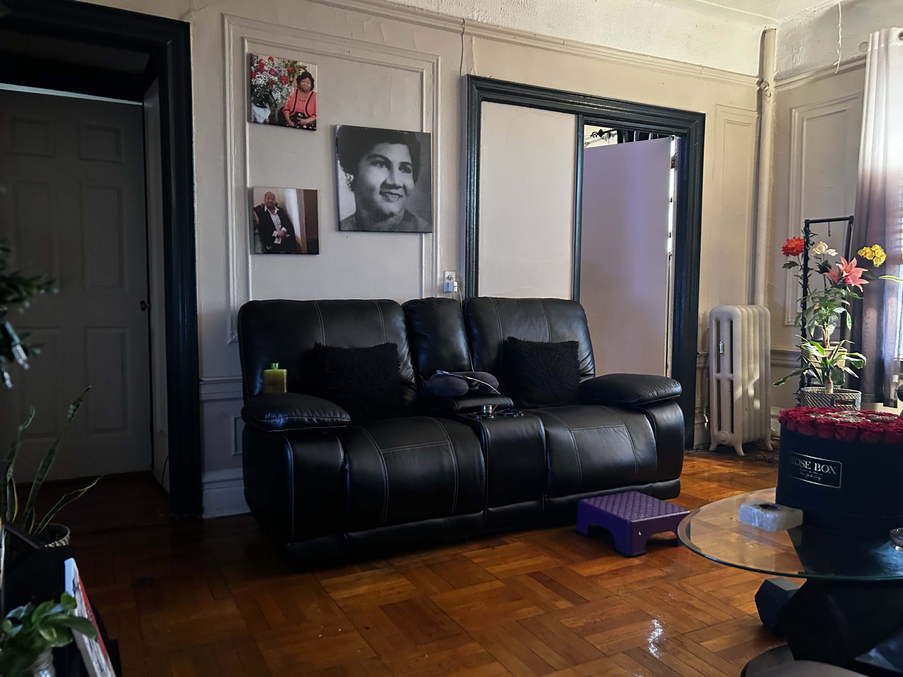 A living room with a black couch, a small purple stool, a glass table, and framed photographs on the wall.