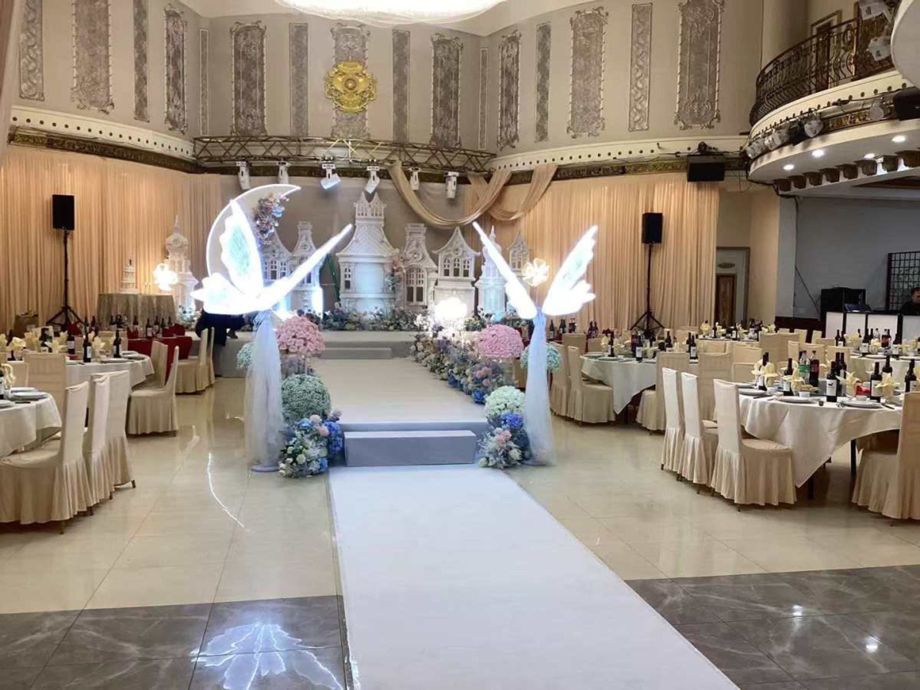 The image shows a beautifully decorated banquet hall with tables set for a formal event, featuring a central pathway adorned with flowers and decorative elements.