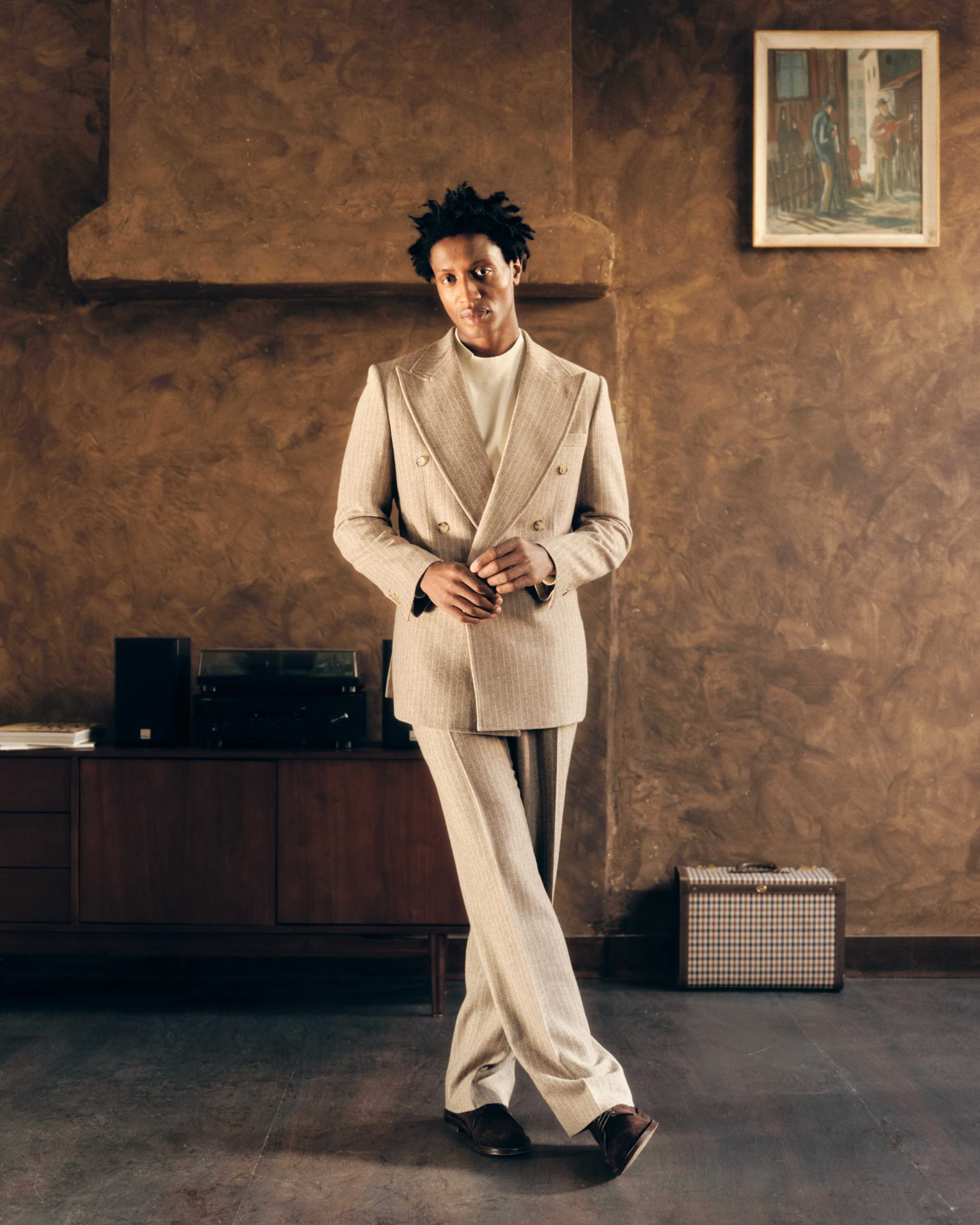 A man wearing a double-breasted tan suit stands in a stylish, dimly-lit room with brown walls and a wooden cabinet.