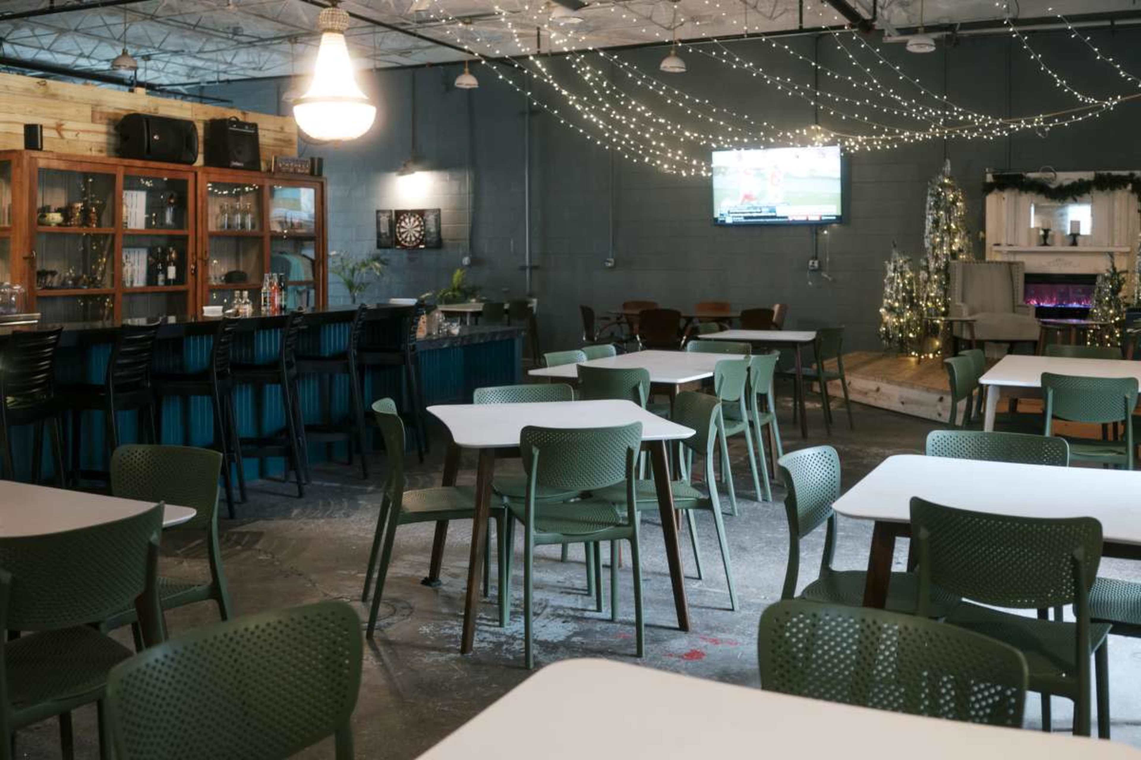 The image shows a spacious interior of a bar or restaurant featuring green chairs and tables, a bar area with shelves, and wall decorations, along with a TV displaying a sports game.