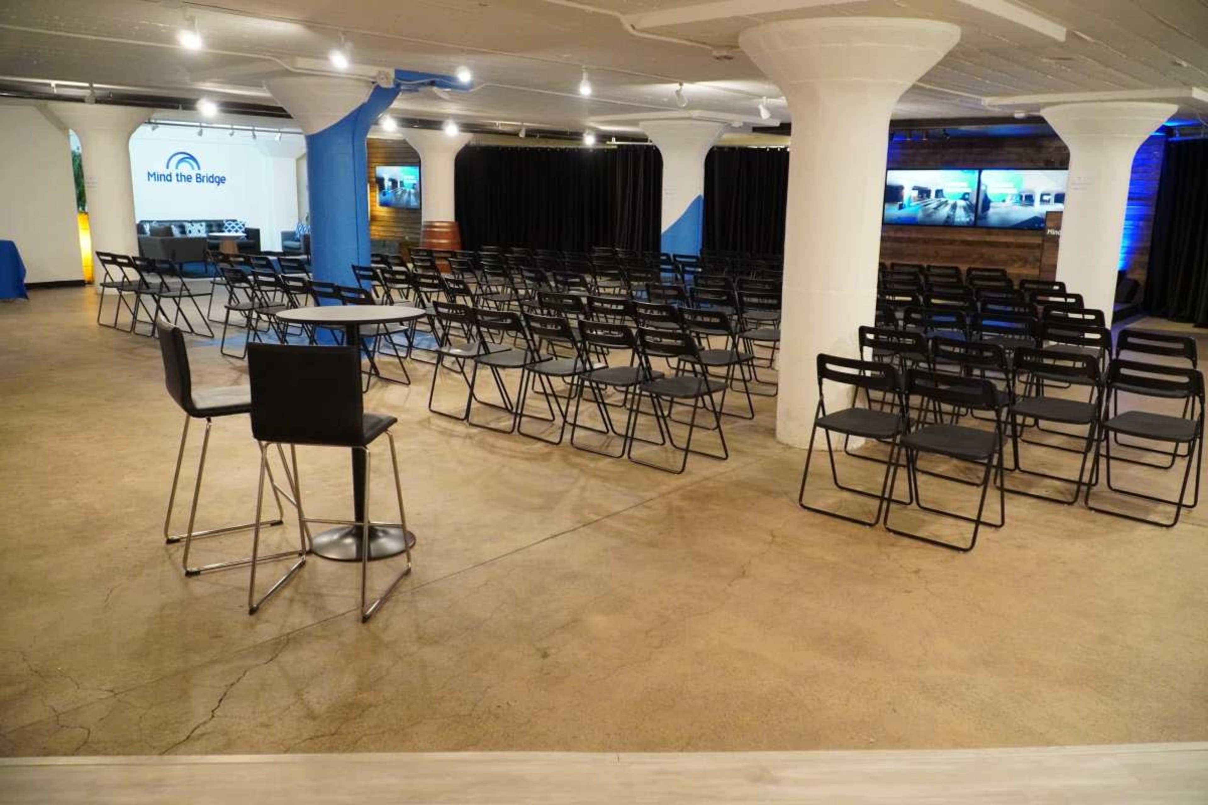A modern event space with rows of black chairs set up in front of a presentation area and a large screen, with white columns and blue accents in the background.