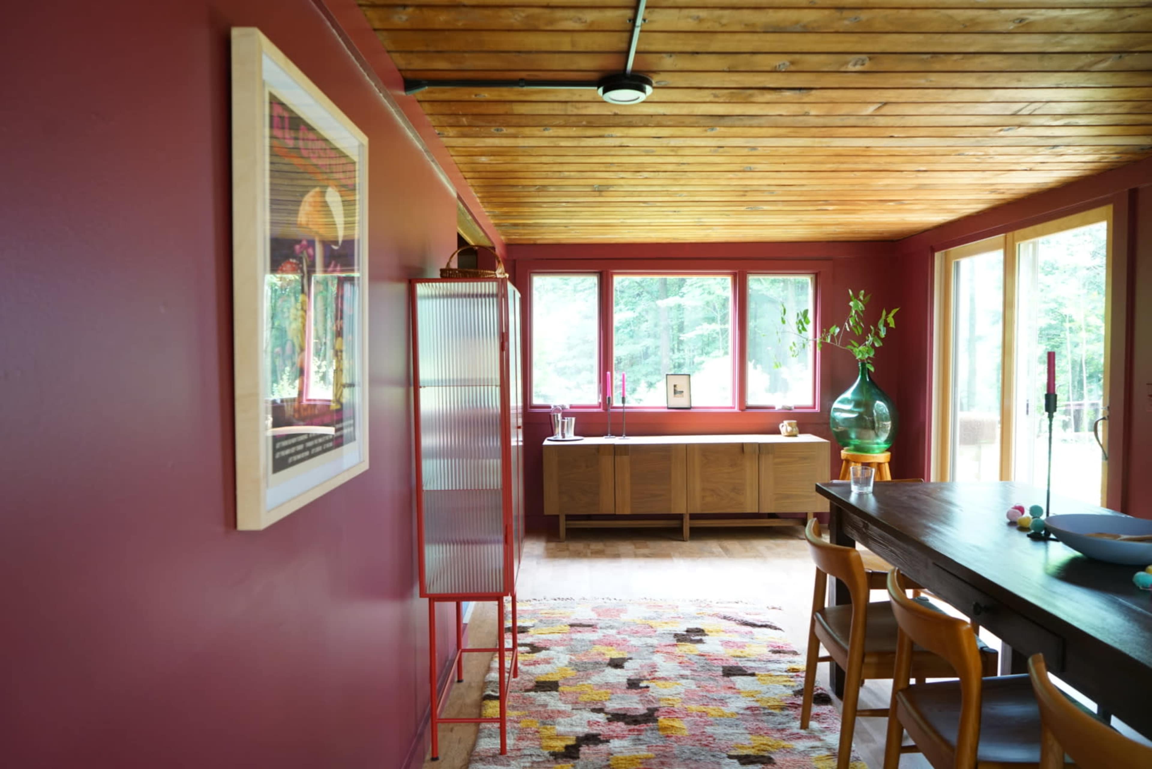 A dining area with a wooden ceiling, red walls, a large table, a sideboard, and several windows allowing natural light.