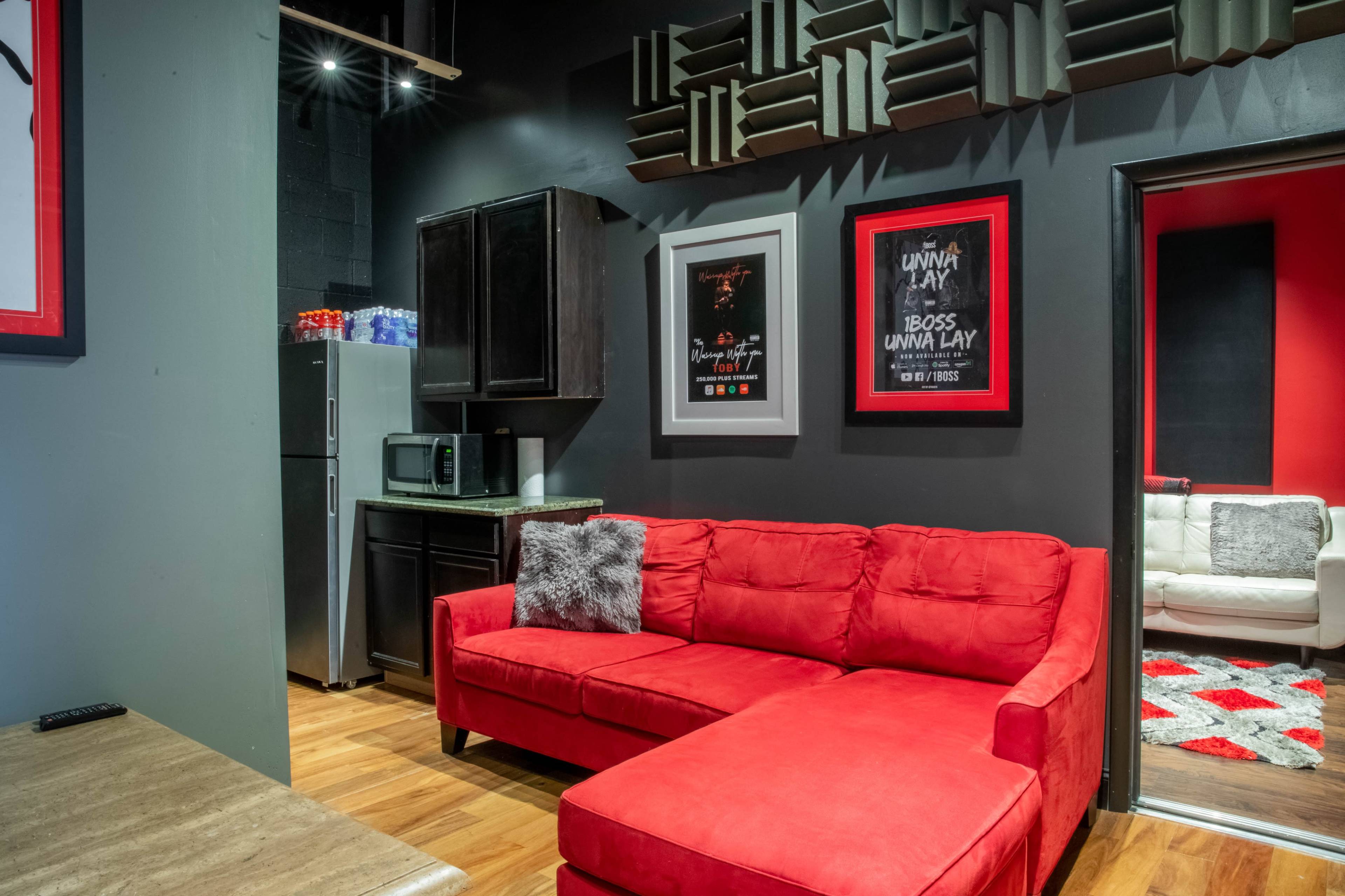 A modern room featuring a red sectional couch, a dark kitchenette with a refrigerator and microwave, and framed artwork on the walls.