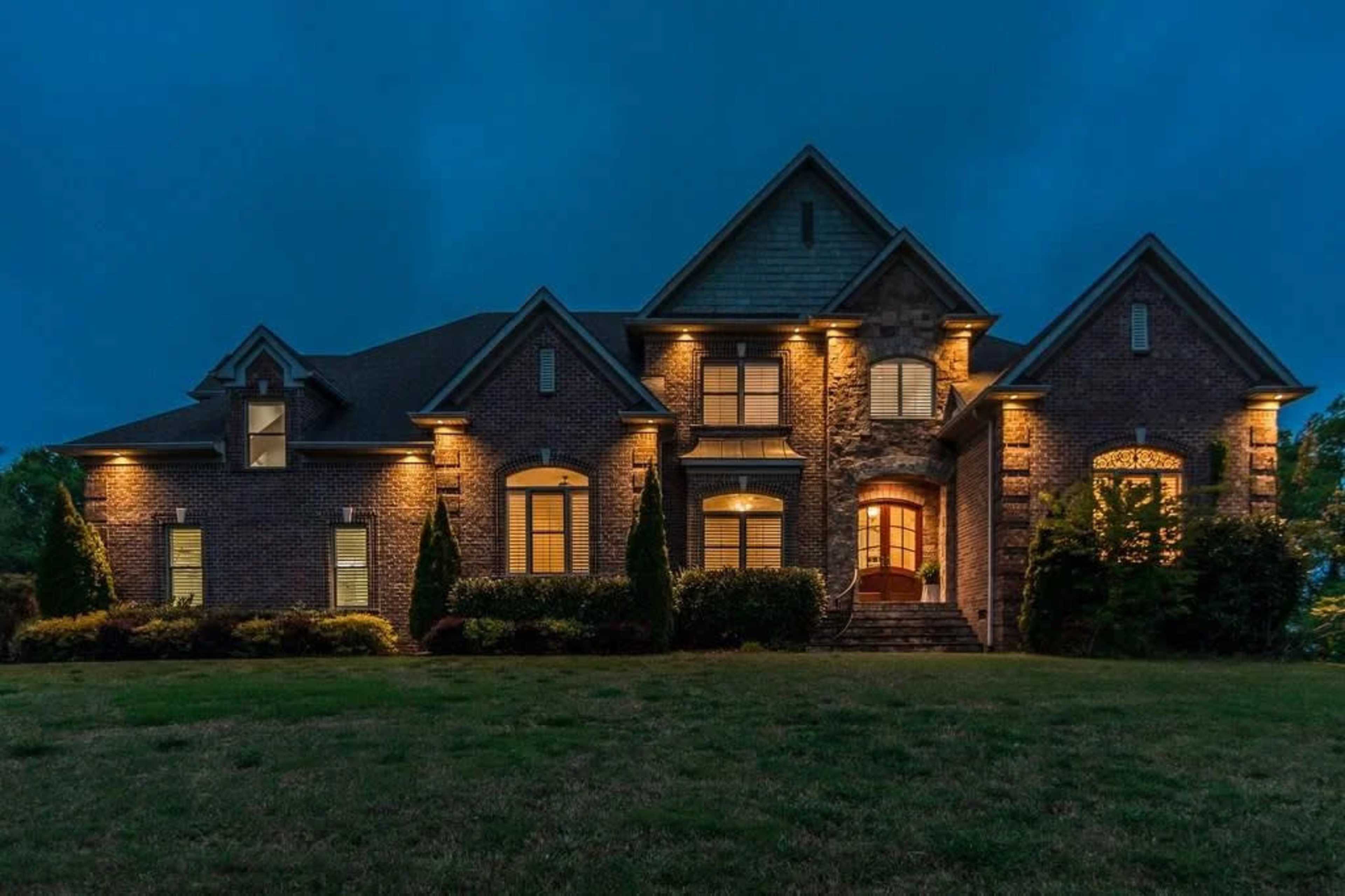 A large brick house with multiple gabled roofs is illuminated by outdoor lights against a twilight sky.