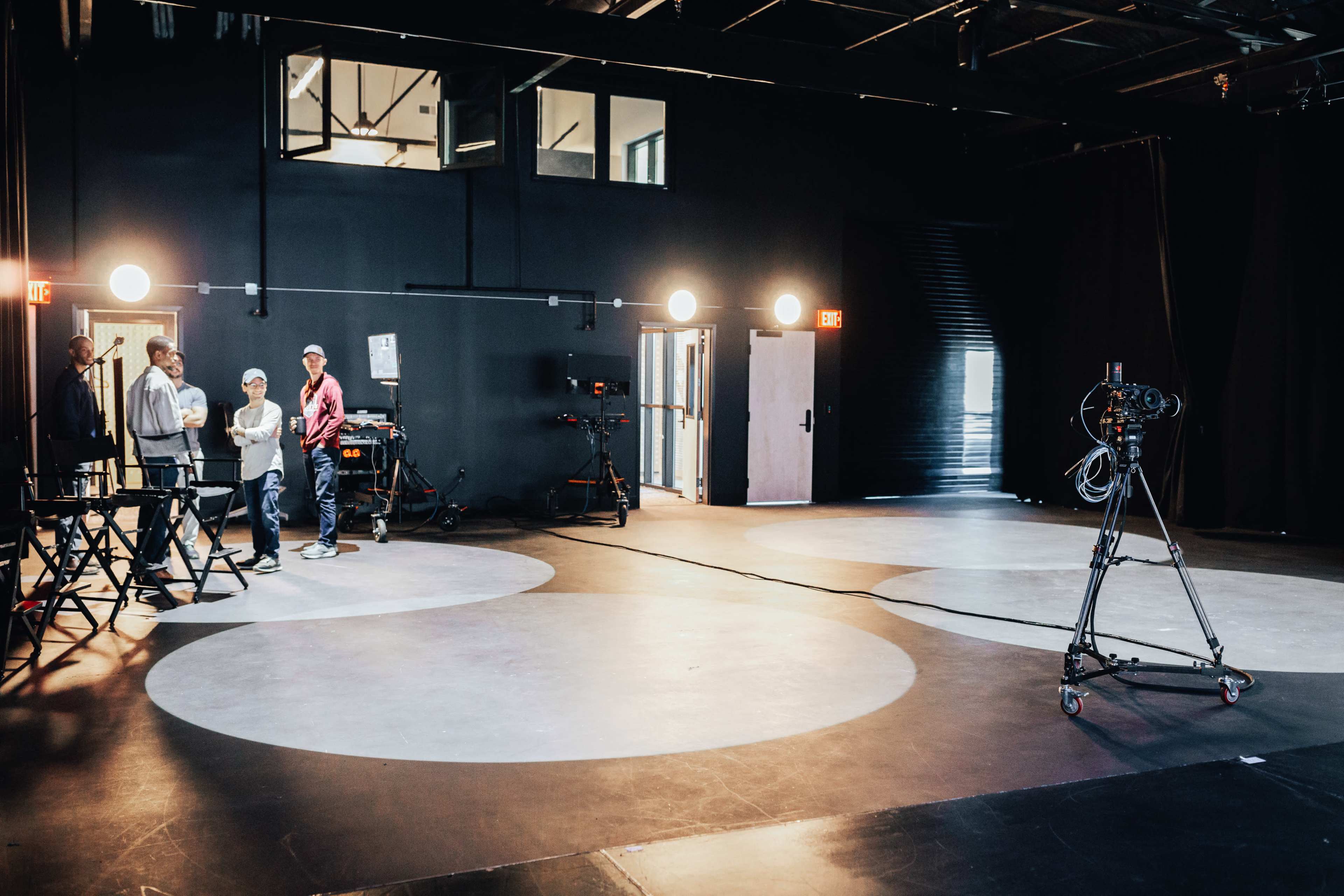 A film studio features circular lit areas on the floor, with equipment and crew members standing nearby.