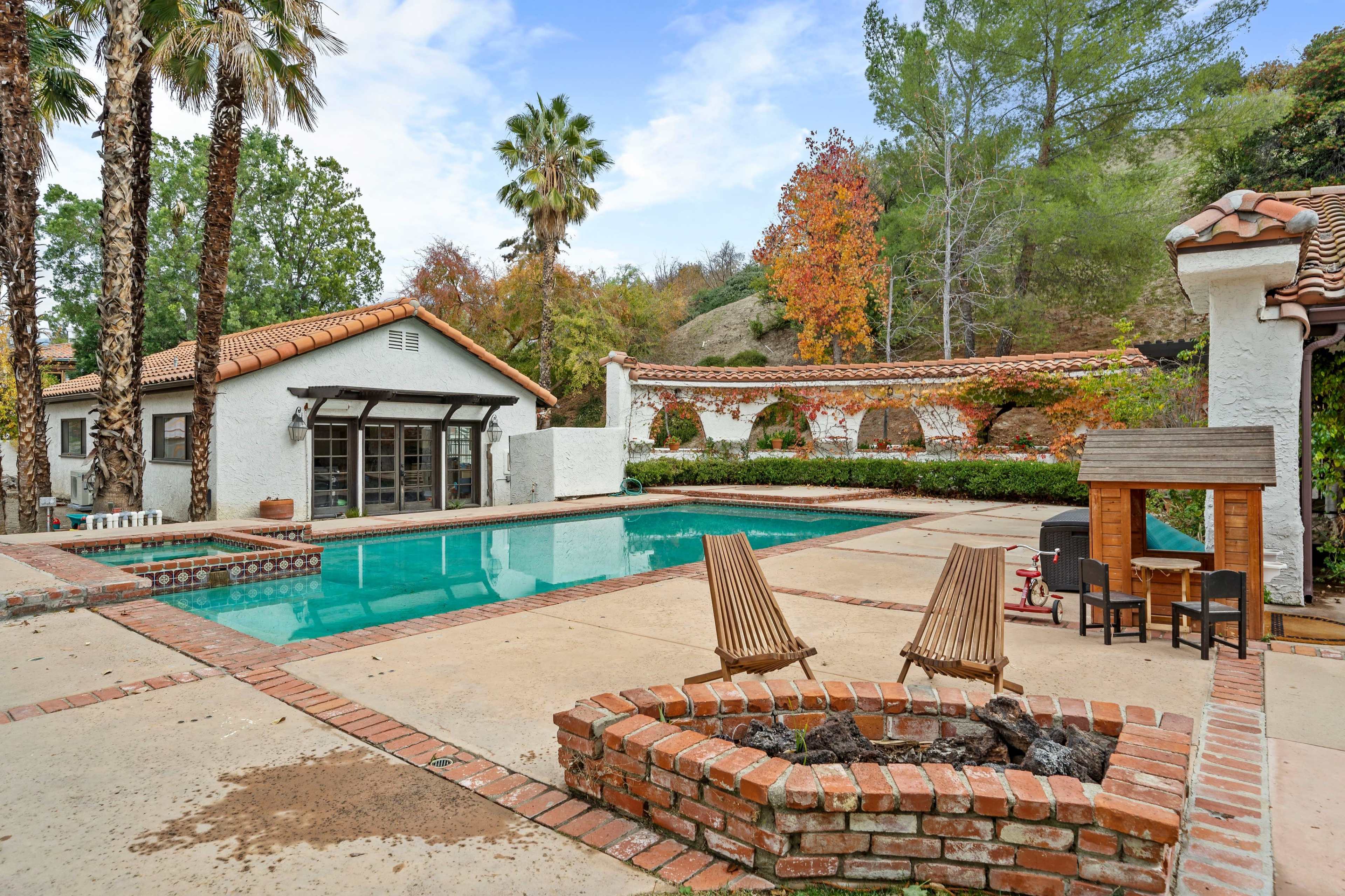 A pool surrounded by landscaping and outdoor seating complements a white stucco house in a wooded area.