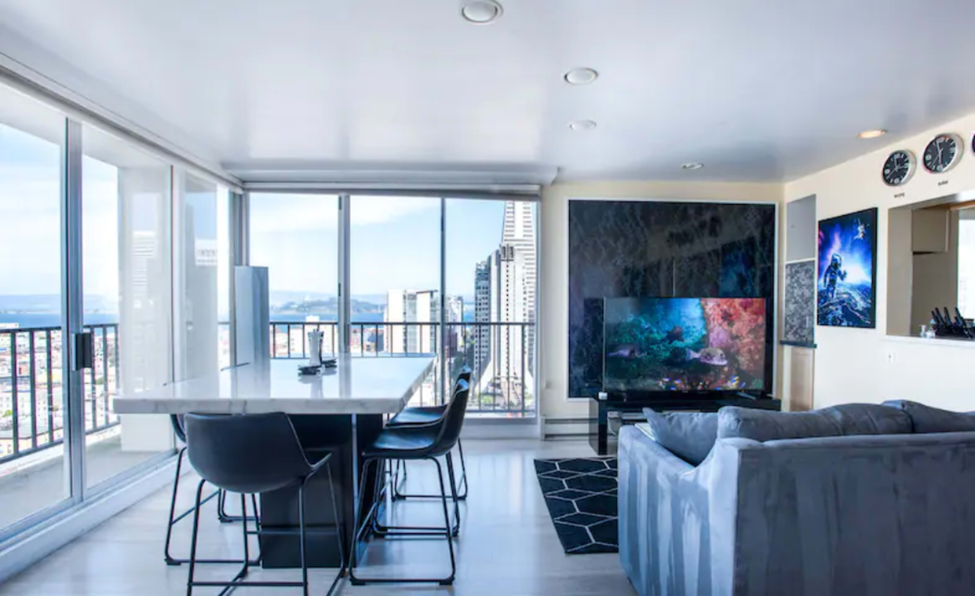 A modern living space features a sleek kitchen island with bar stools, a large television, and expansive windows showcasing a city view.