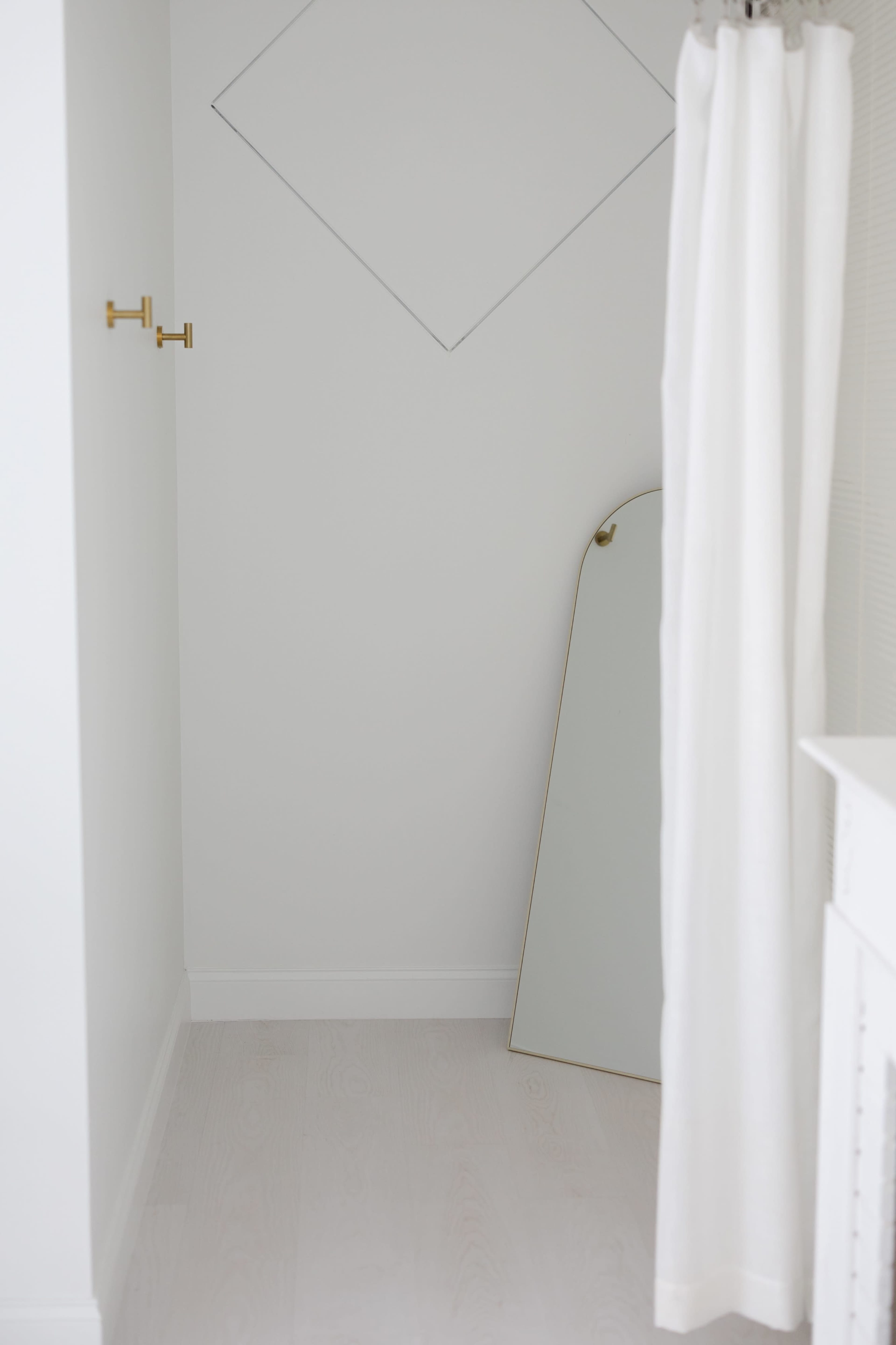 The image shows a minimalistic corner of a room featuring a large mirror leaning against the wall, white curtains, and two gold hooks.