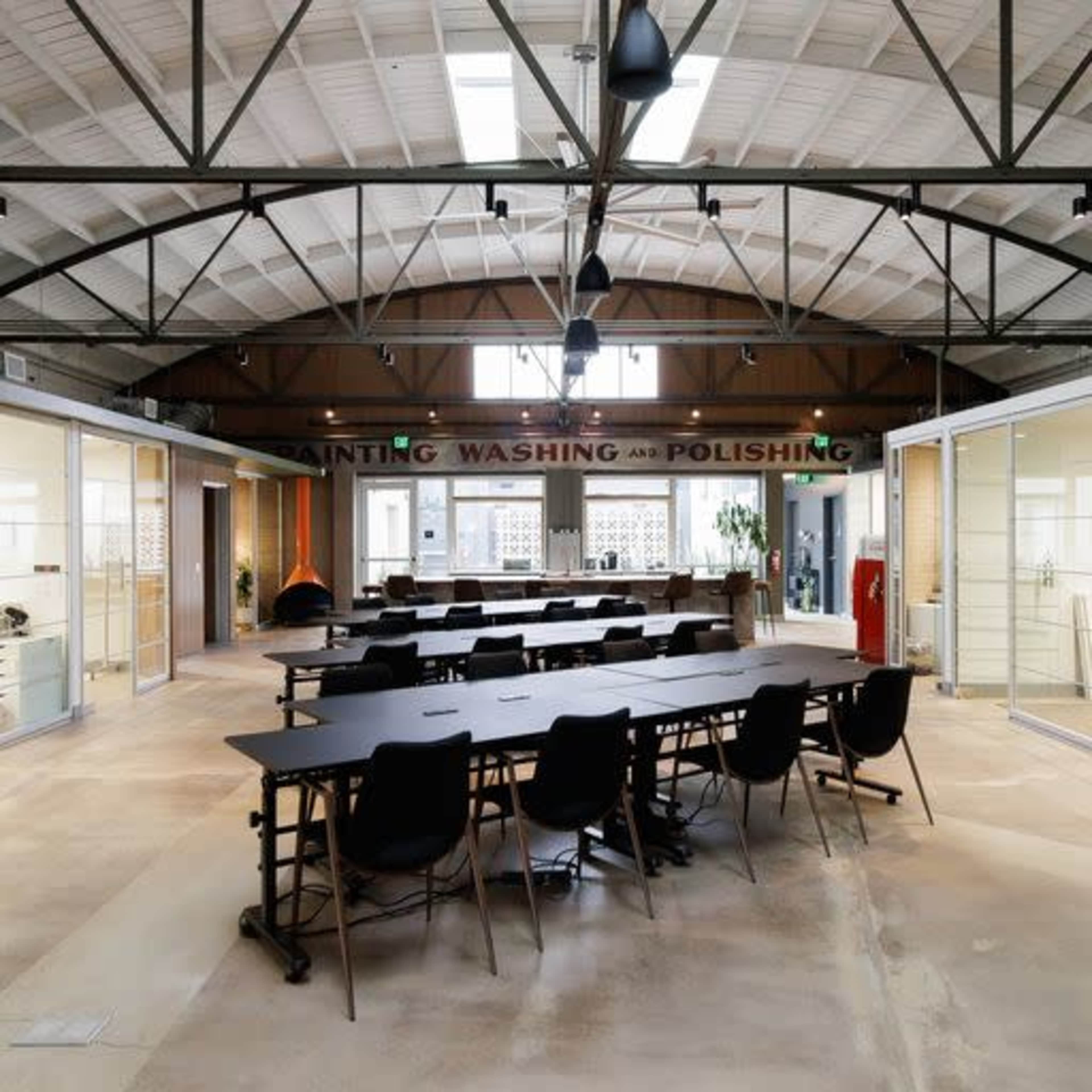 A spacious industrial workspace with a long table and several black chairs, surrounded by glass-walled offices and signs indicating areas for painting, washing, and polishing.