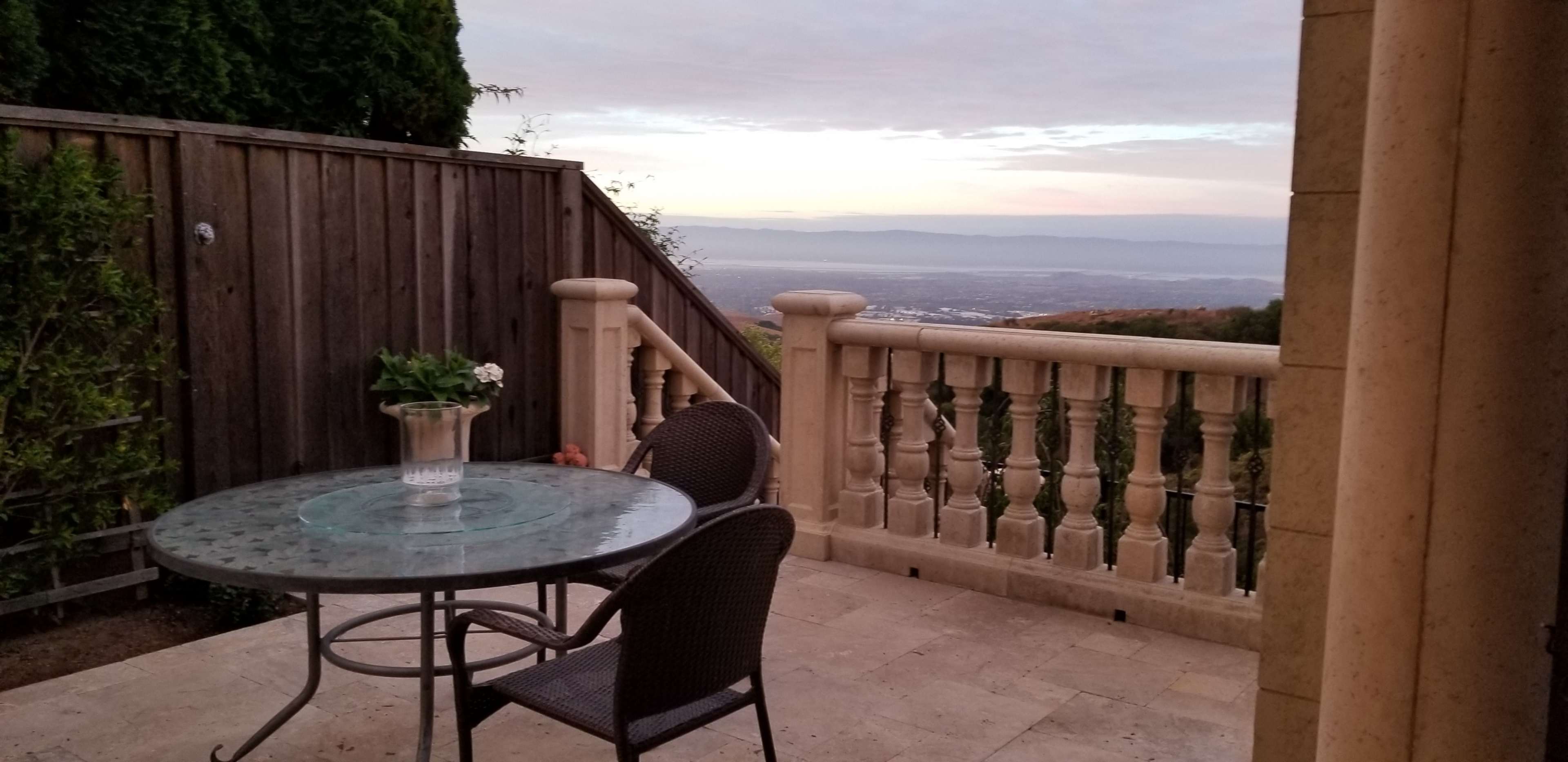 A patio with a glass-top table and two chairs overlooks a valley at sunset, framed by a wooden railing and stone columns.