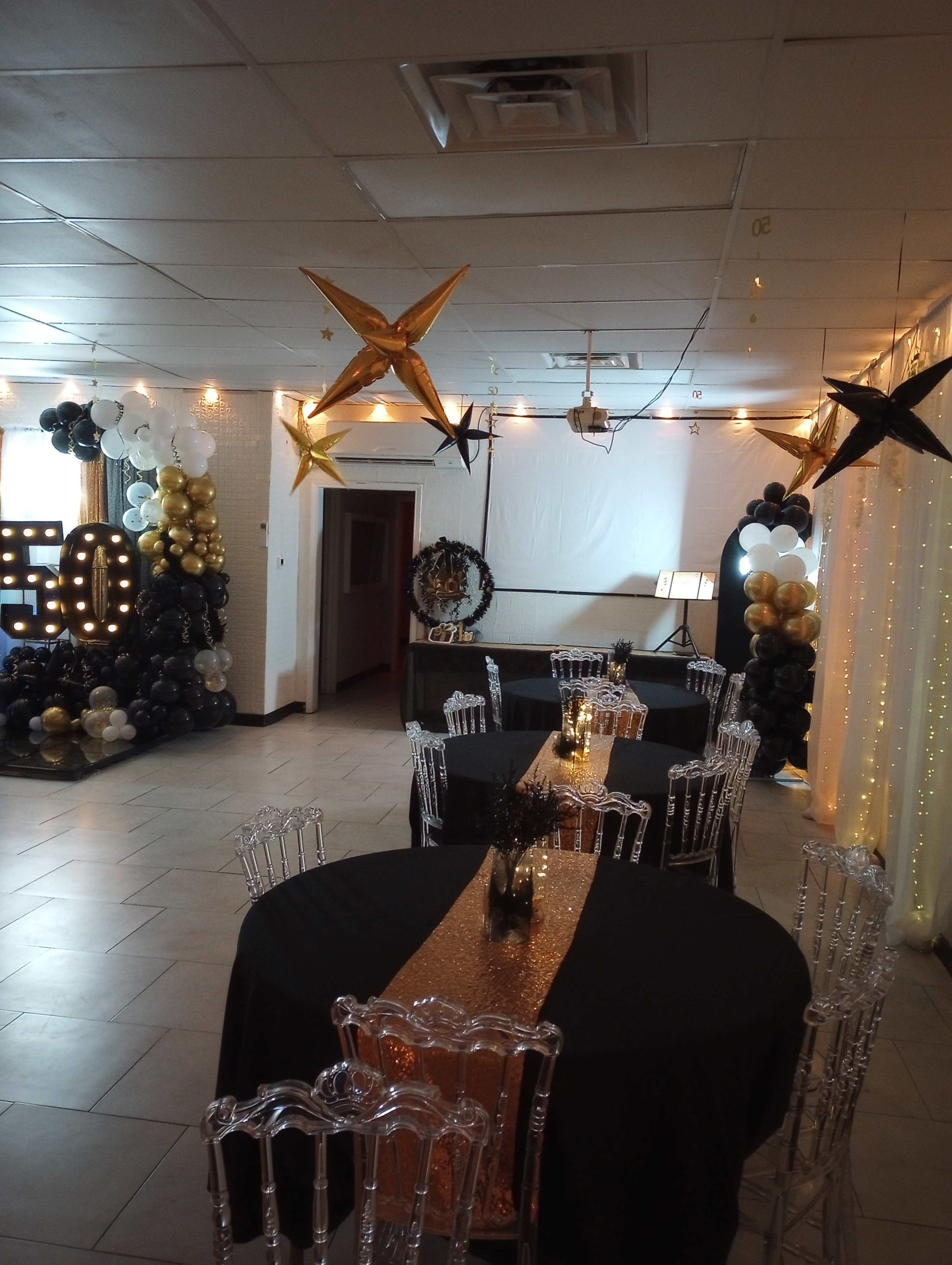 The interior of a decorated event space features black tables with gold runners, clear chairs, and festive balloon arrangements in gold, black, and white.