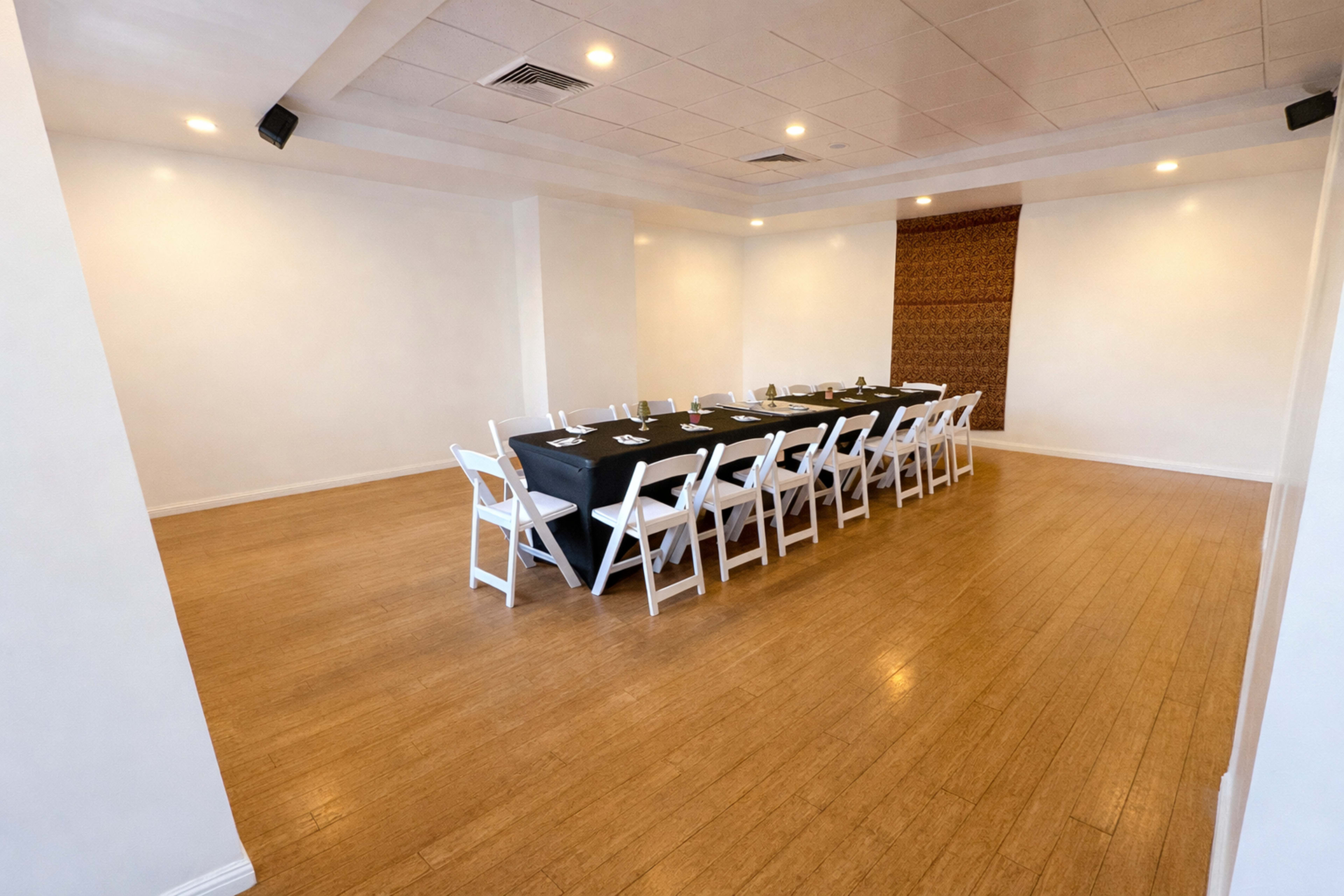 A long table with black linens and white chairs is set up in a spacious, well-lit room with plain white walls and a decorative feature on one side.