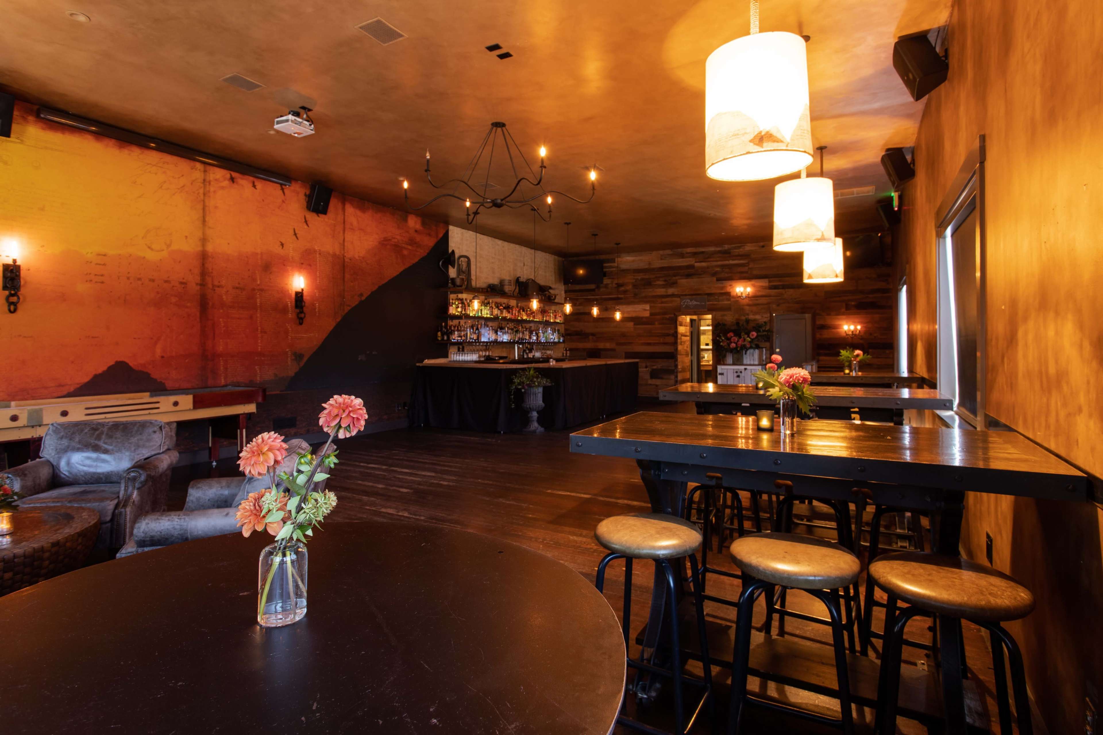 A dimly lit bar area with wooden walls, tables with vases of flowers, and a shelf stocked with bottles in the background.
