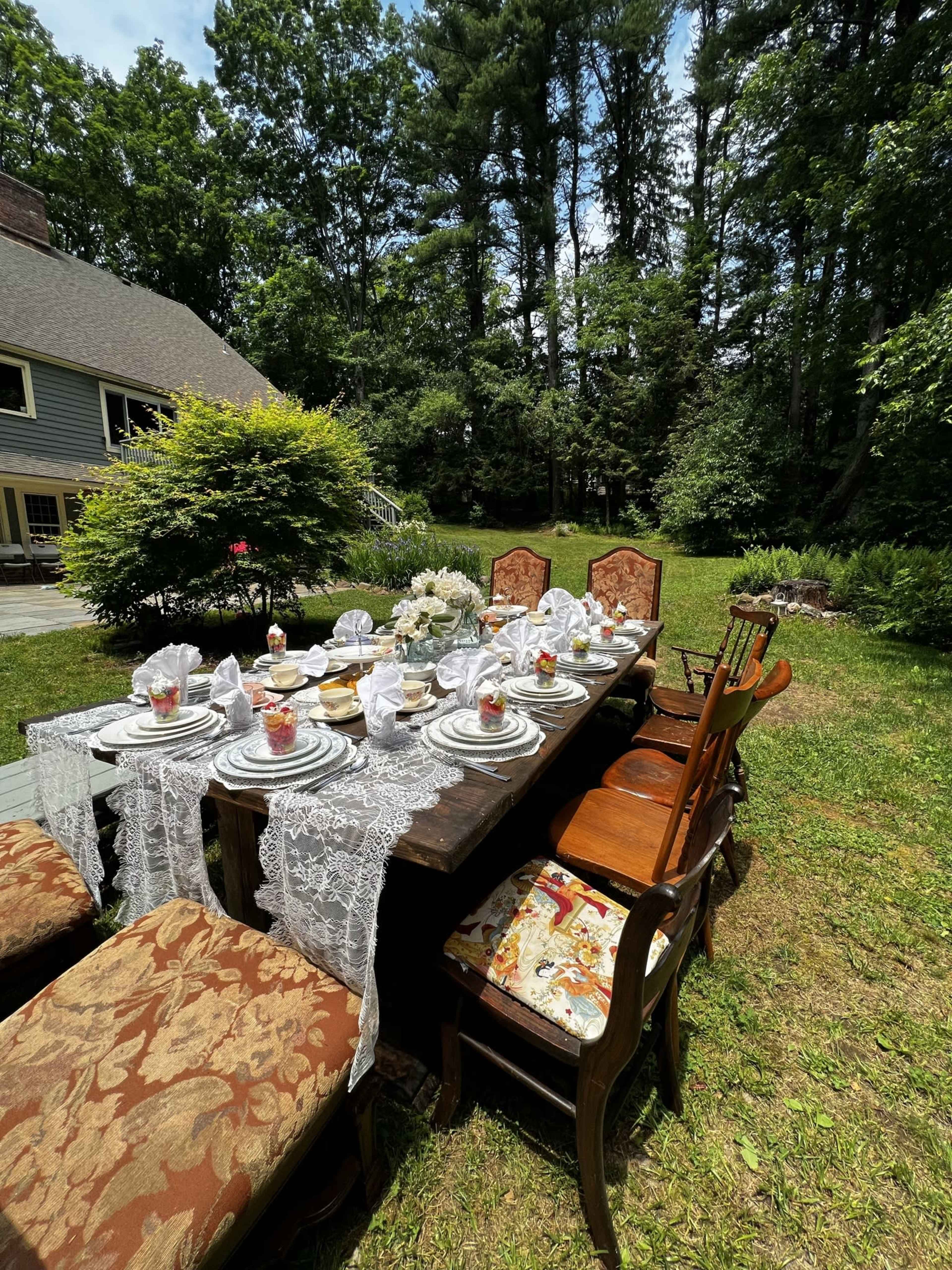 A long outdoor dining table is set with dishes, glasses, and decorative napkins, surrounded by chairs in a forested backyard.