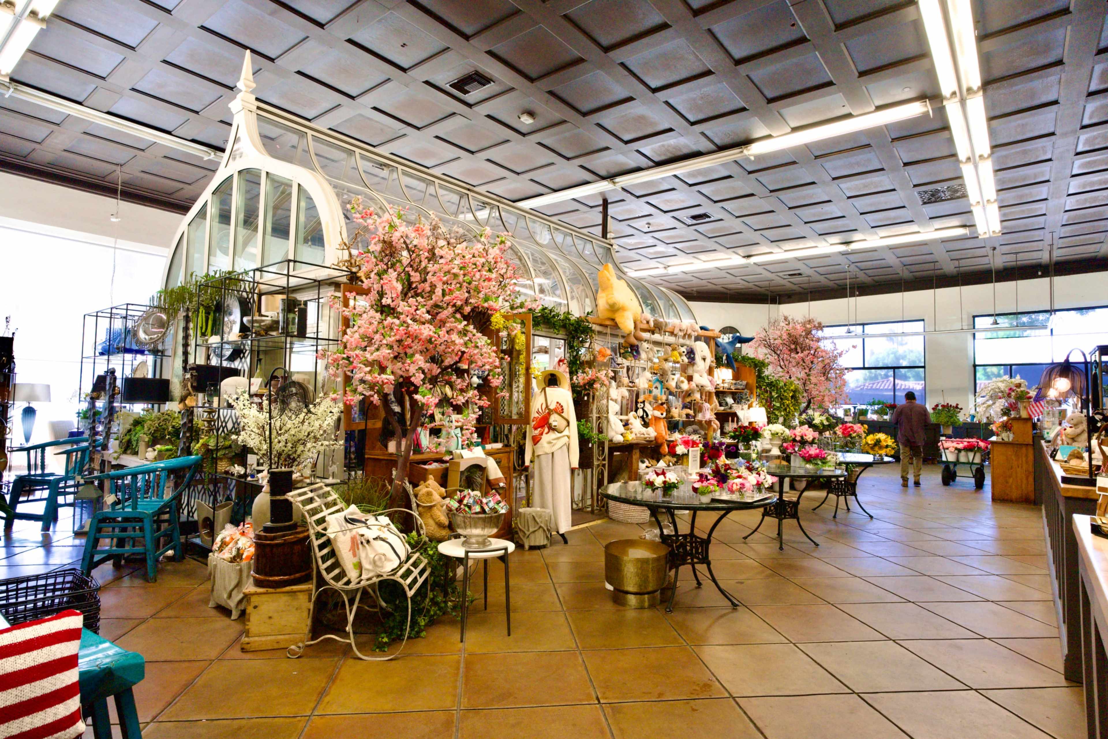 The image shows a brightly lit floral shop featuring an array of plants, flowers, and gardening accessories, with a large greenhouse-like structure at its center.