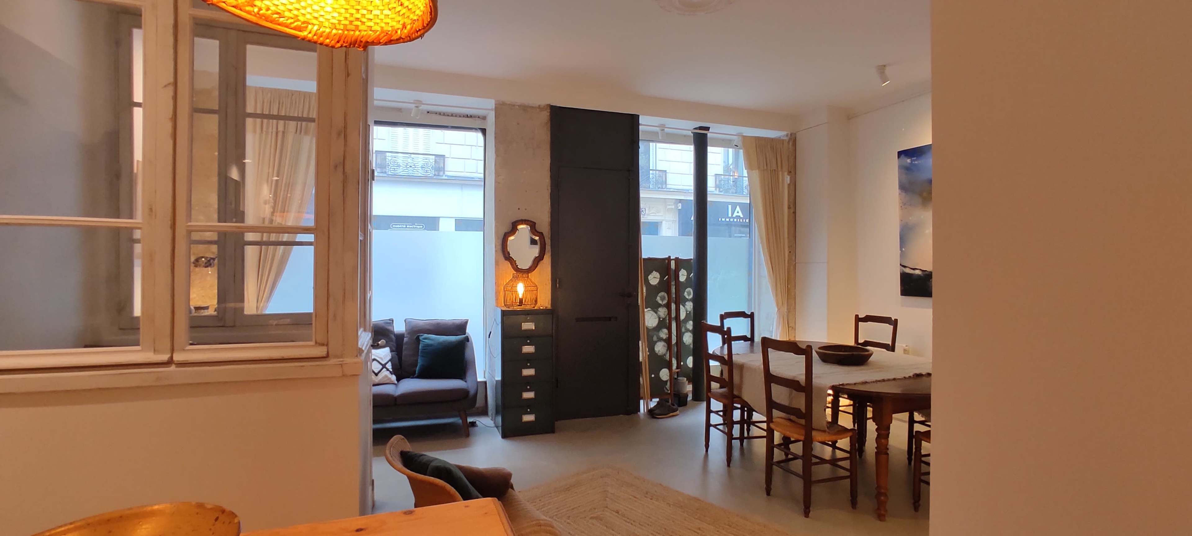 The image shows a modern living space featuring a dining area with wooden furniture, a cozy seating area with a couch, and large windows allowing natural light to enter.
