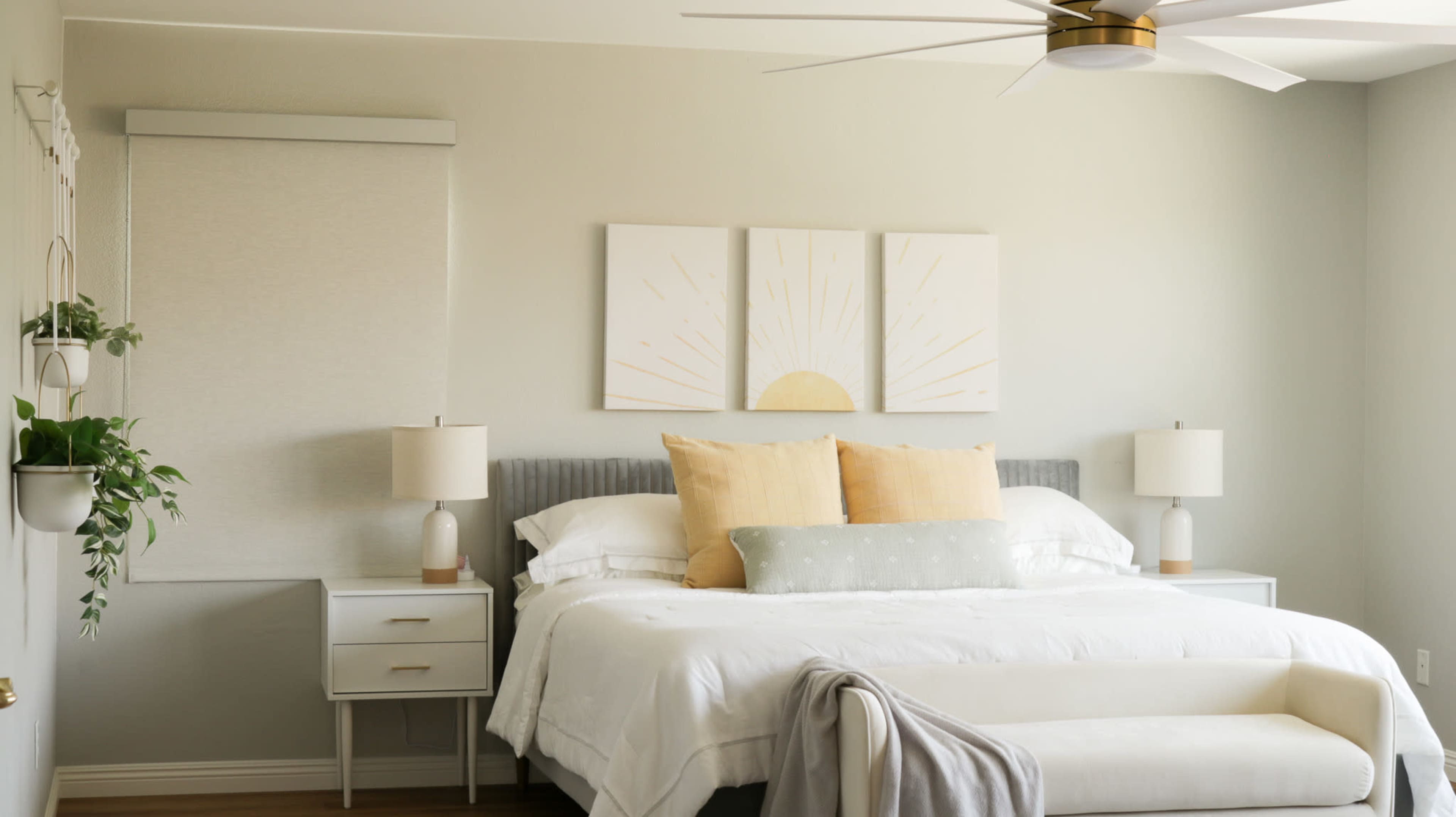 A light-colored bedroom features a bed with white linens and yellow accent pillows, flanked by two bedside lamps and adorned with three sunburst paintings above the headboard.