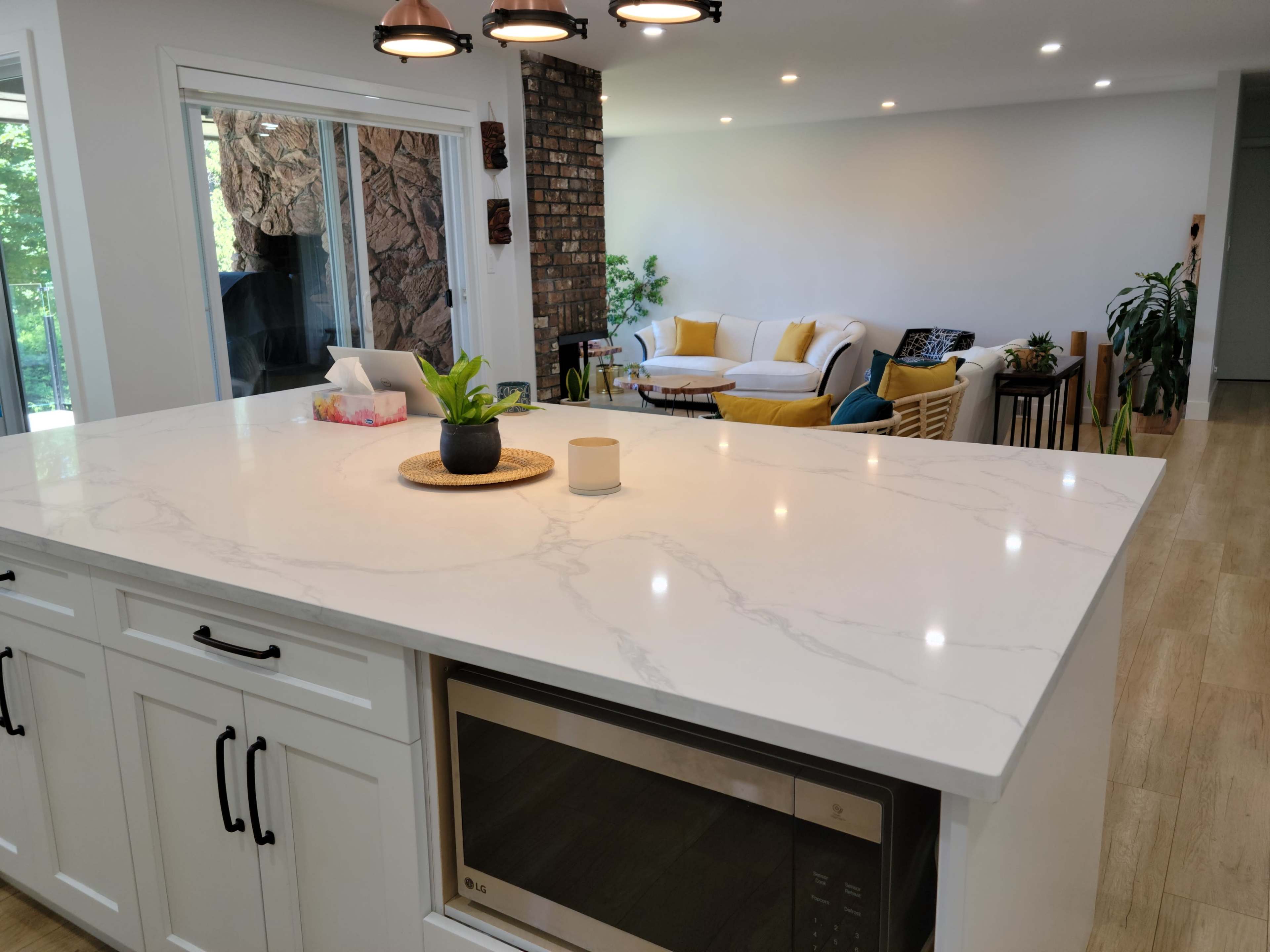 A modern kitchen with a marble countertop, featuring a microwave on the lower cabinet, overlooks a living area with a sofa and plants.