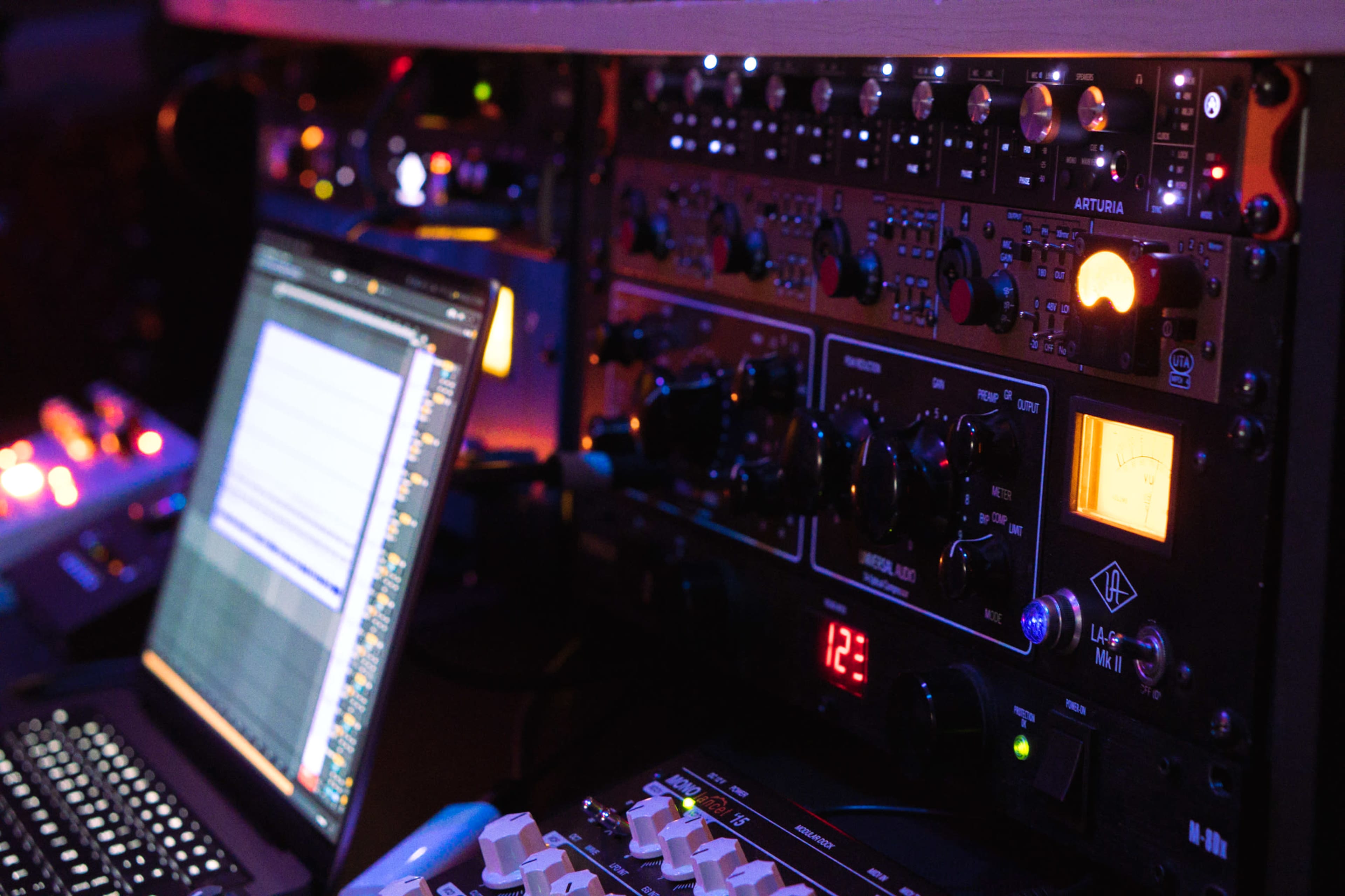 The image shows a close-up of audio equipment, including mixers and a computer screen, in a dimly lit studio setting.