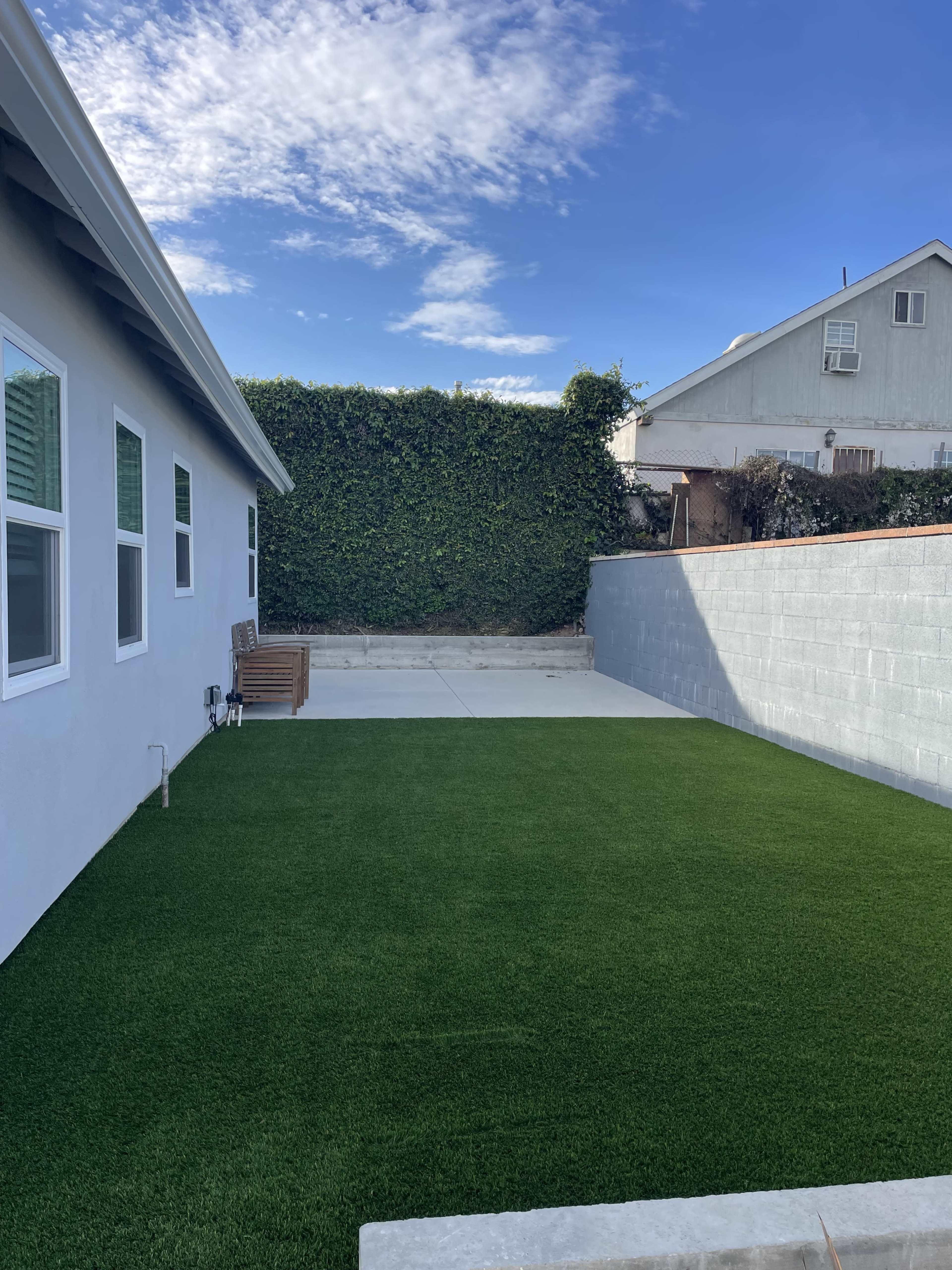 The image shows a landscaped backyard featuring artificial turf, a concrete patio, and a wall covered with greenery.