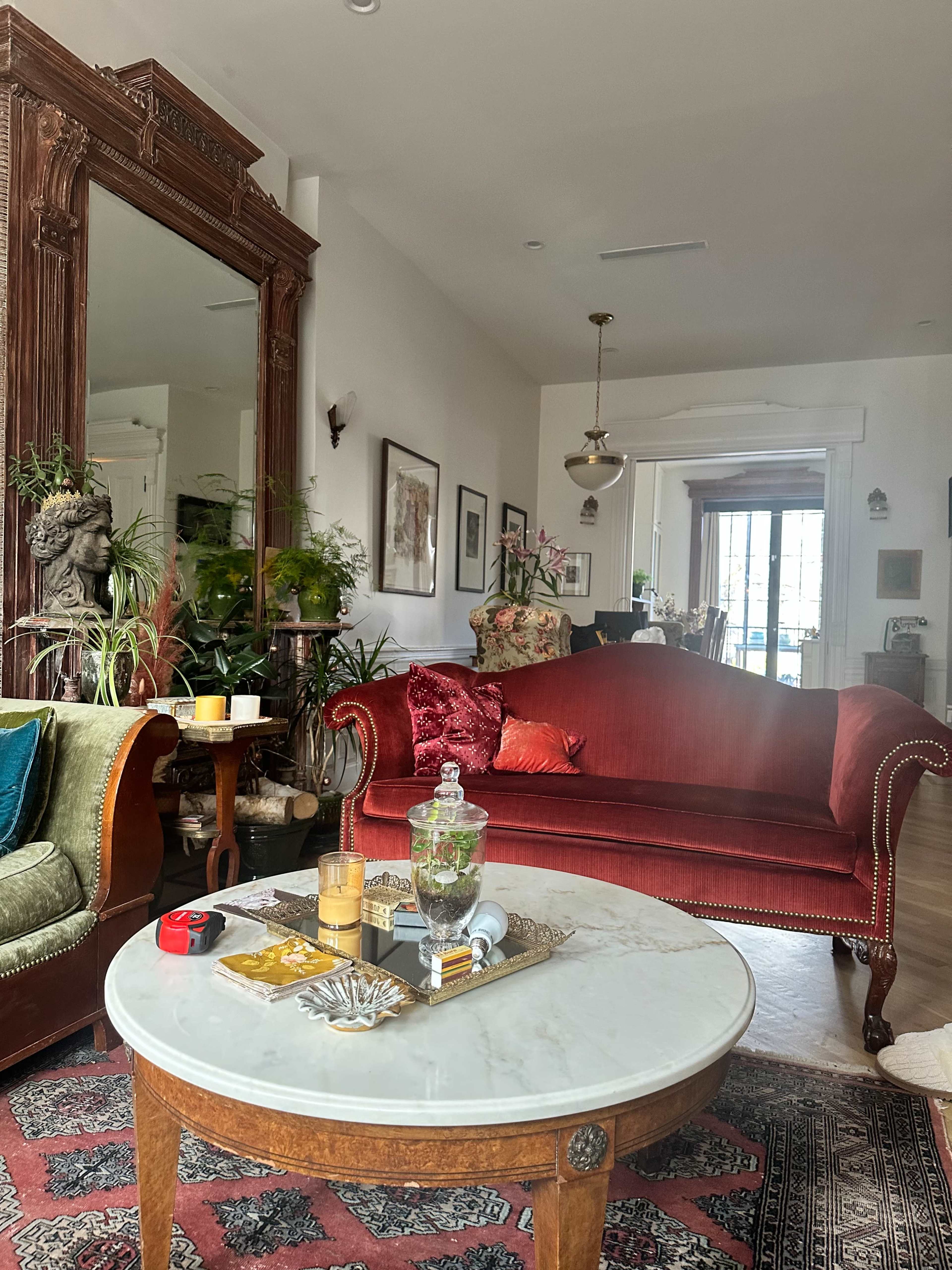 The living room features a red velvet sofa, a marble coffee table with decorative items, and various plants, framed artwork, and a large ornate mirror.