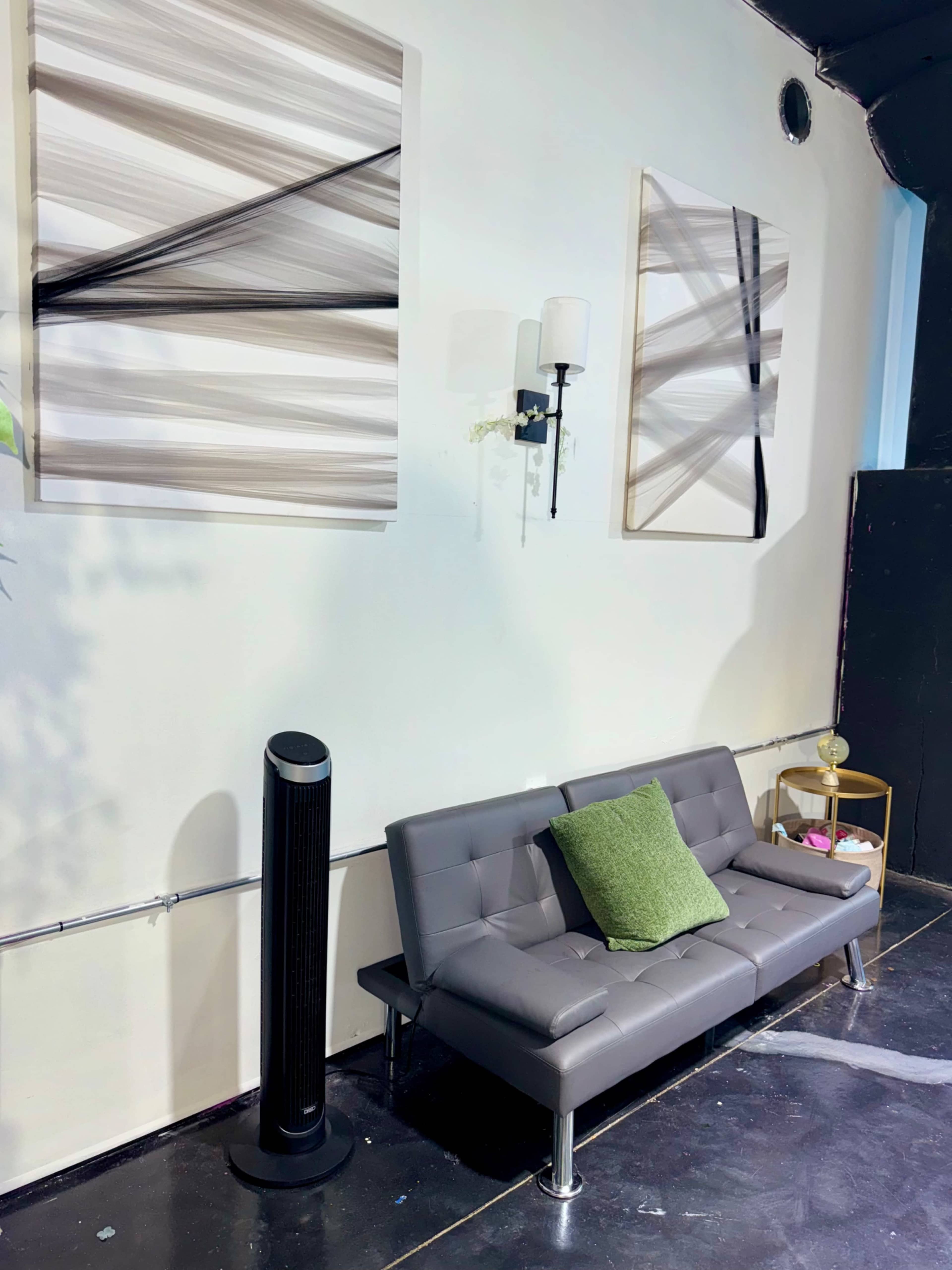 The image shows a modern gray couch with a green throw pillow, a tall black fan, and a small round table with items on it, all set against a white wall featuring two abstract artworks.