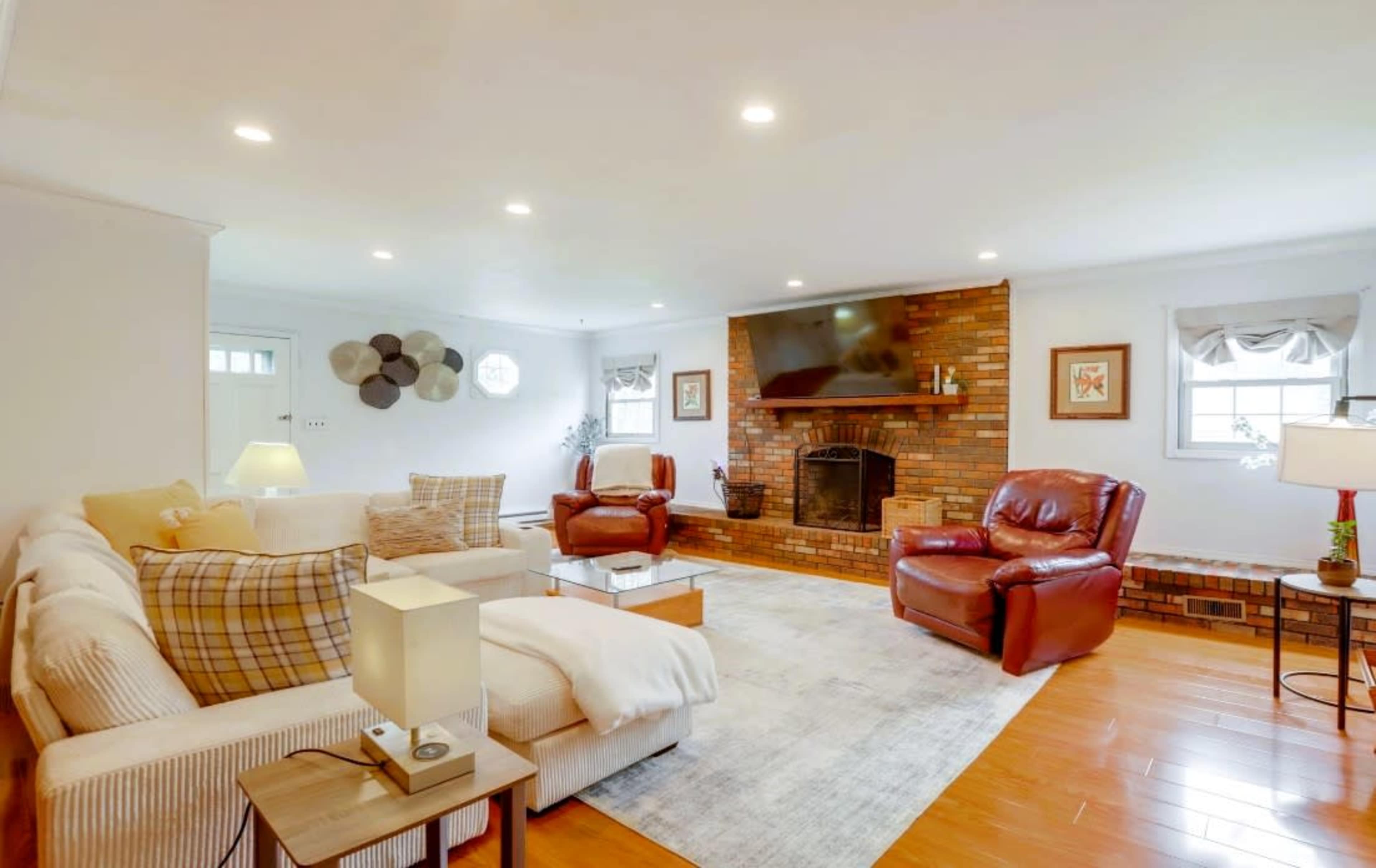 A spacious living room features a brick fireplace with a television above, surrounded by beige sofas and red leather armchairs on a wooden floor.