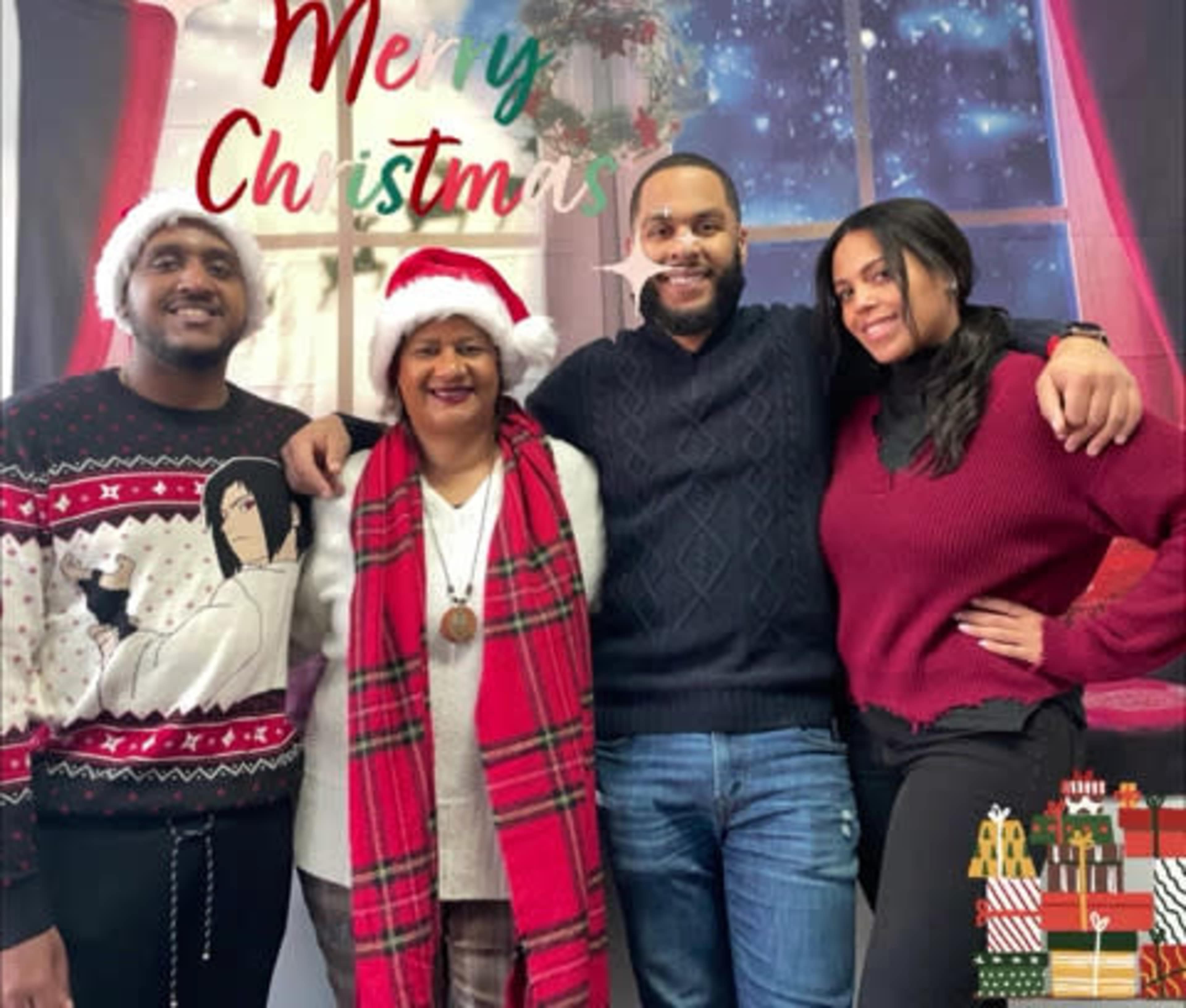 A group of four people wearing festive clothing poses together in front of a holiday-themed backdrop with a Christmas greeting.