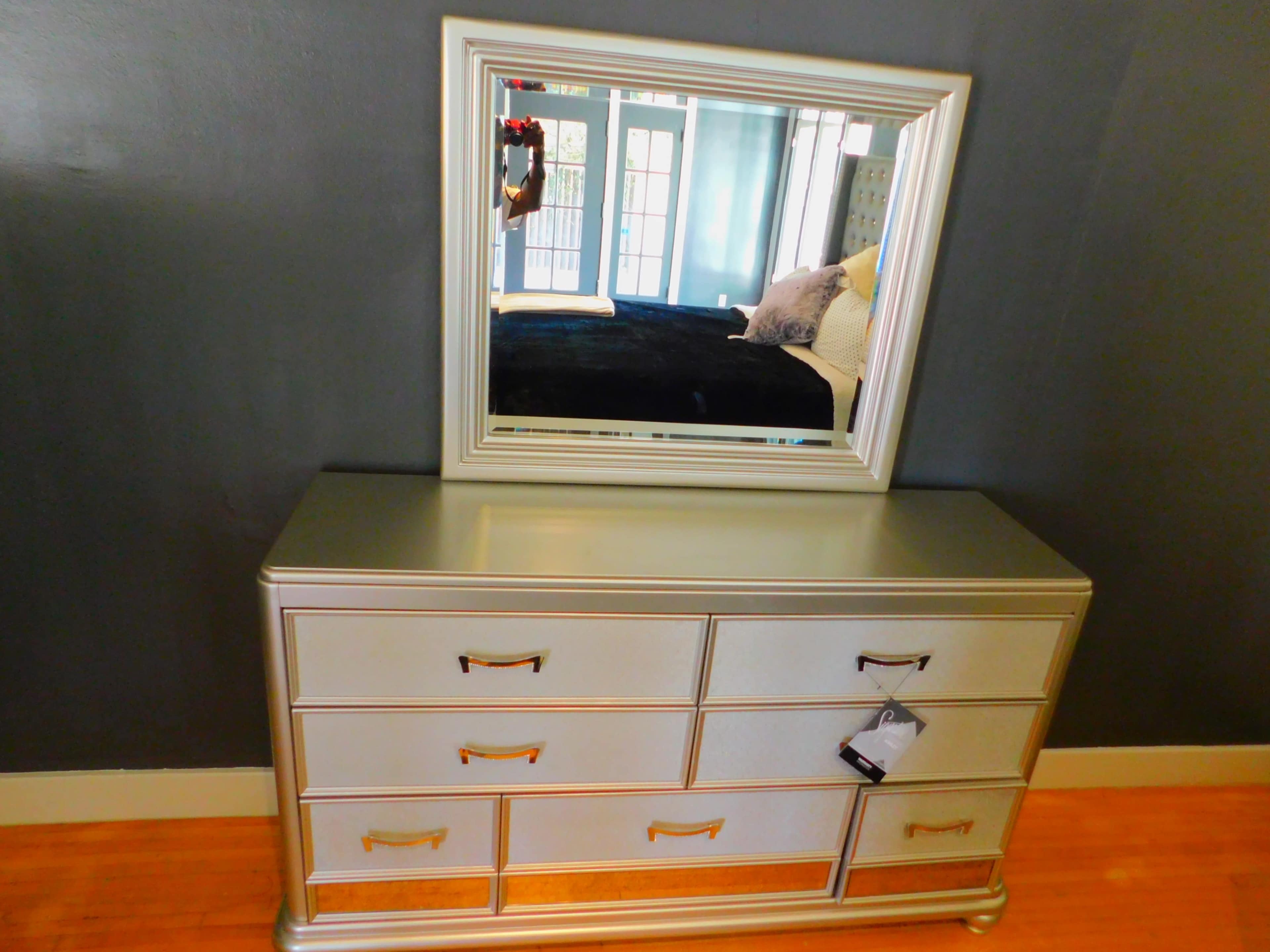 The image shows a silver dresser with multiple drawers and a rectangular mirror mounted above it against a dark wall.