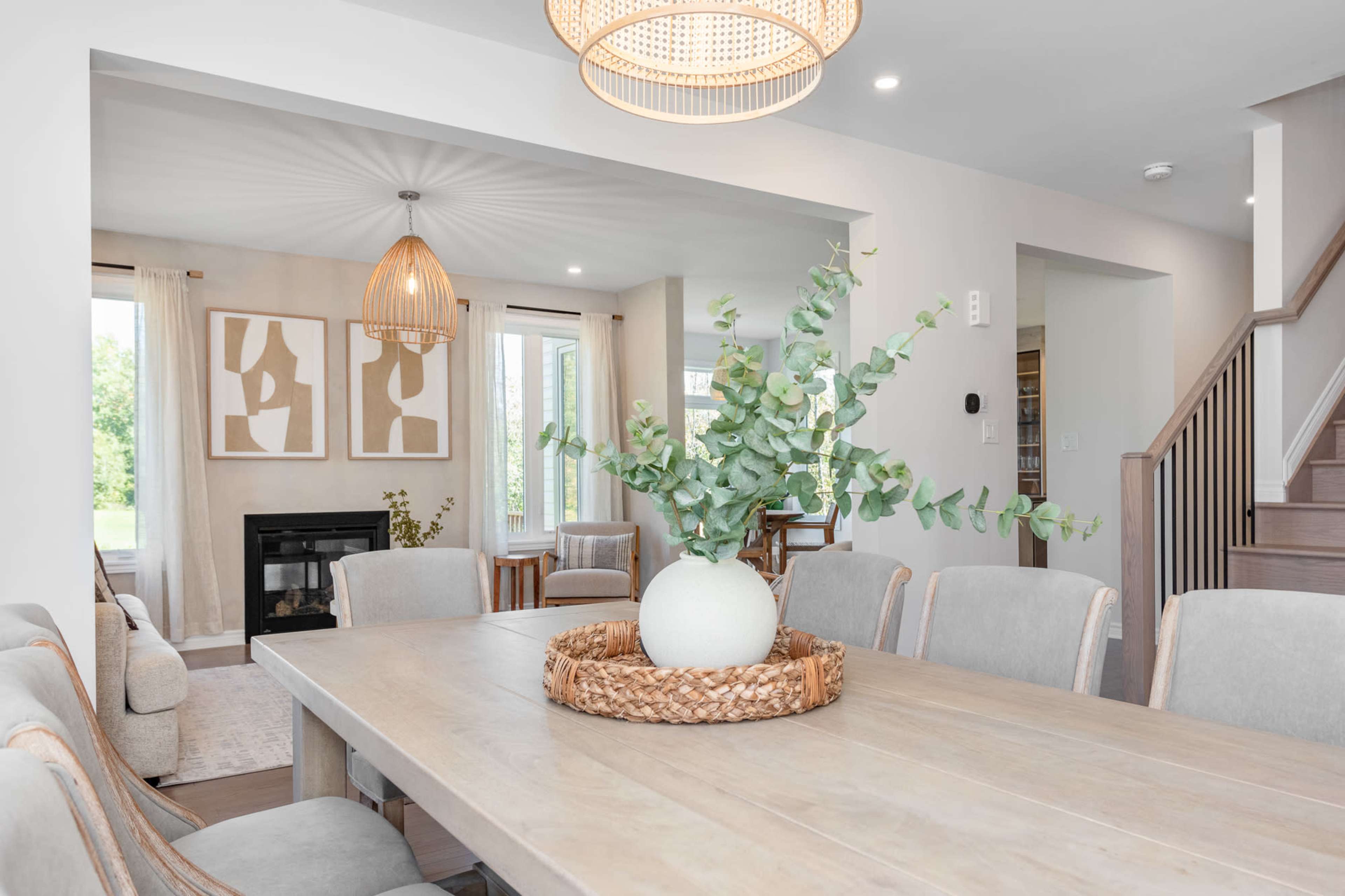 A modern dining room features a light wooden table surrounded by upholstered chairs, with a vase of greenery as the centerpiece and artwork on the walls.