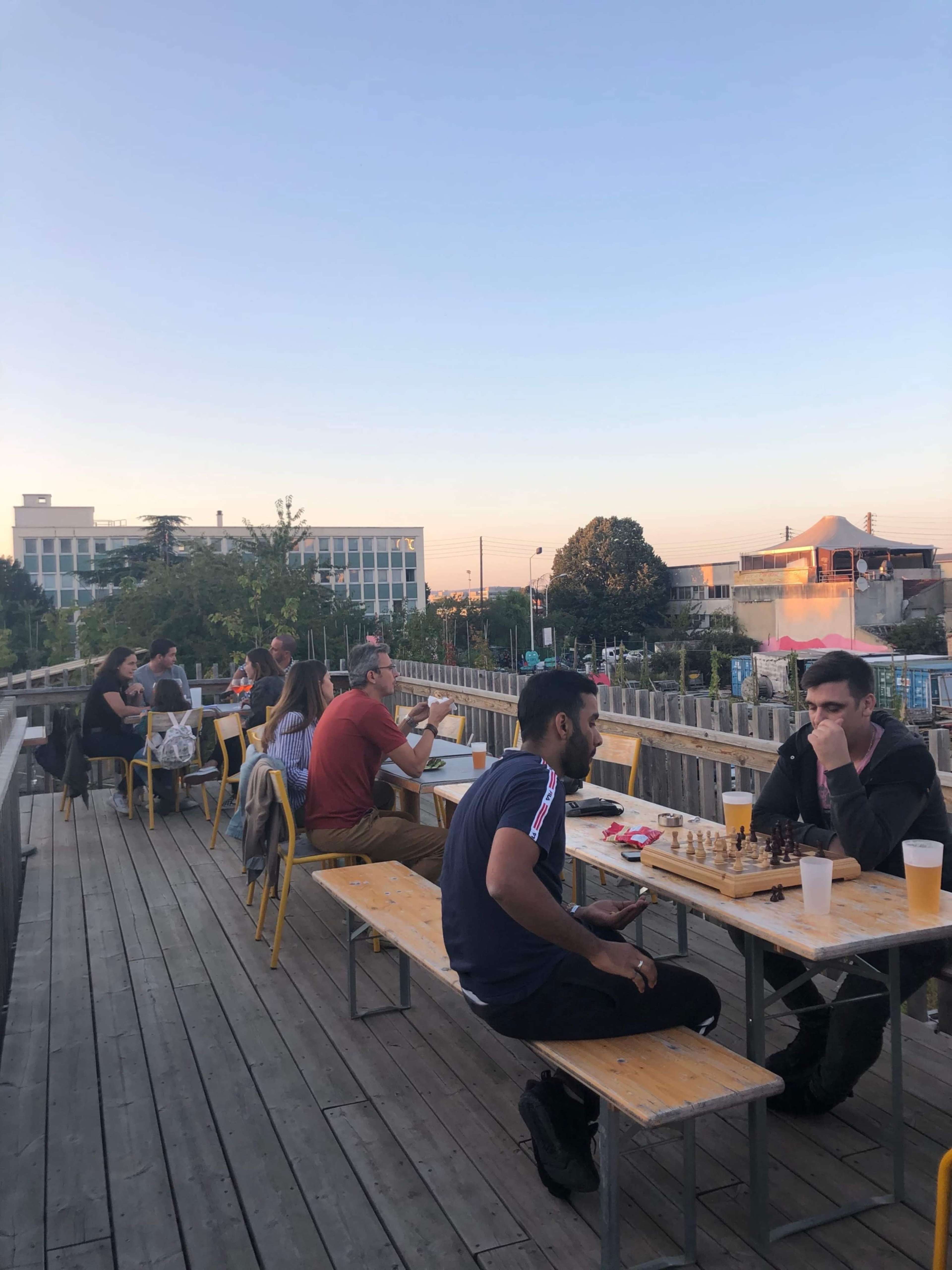 People sit at wooden tables on an outdoor deck, playing chess and enjoying drinks as the sun sets in the background.