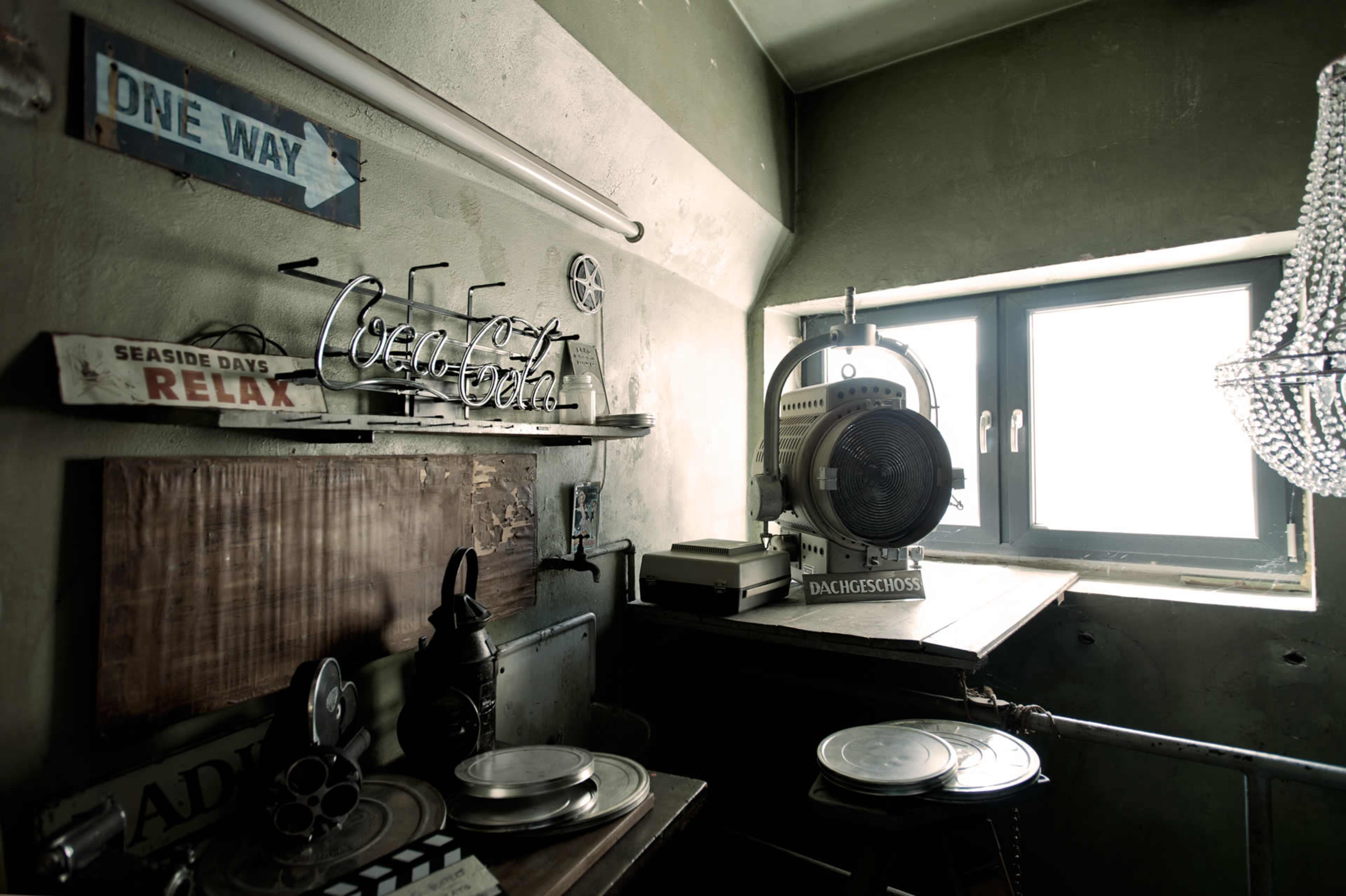 The image shows a cluttered corner of a room featuring vintage signs, a film projector, and a window with light streaming through.