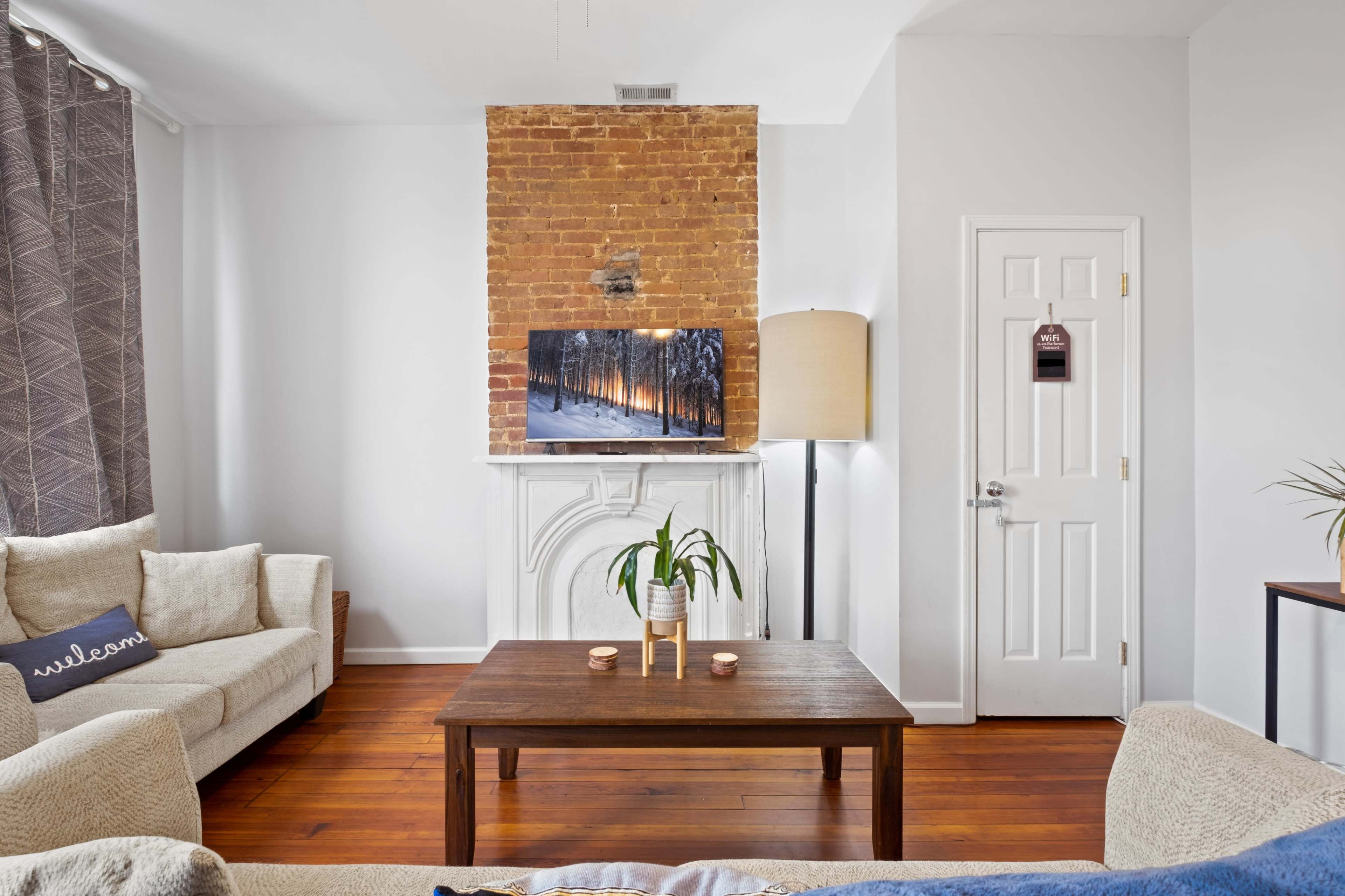 A living room with a brick fireplace, a television mounted above it, a wooden coffee table in the center, and two light-colored sofas positioned around the table.