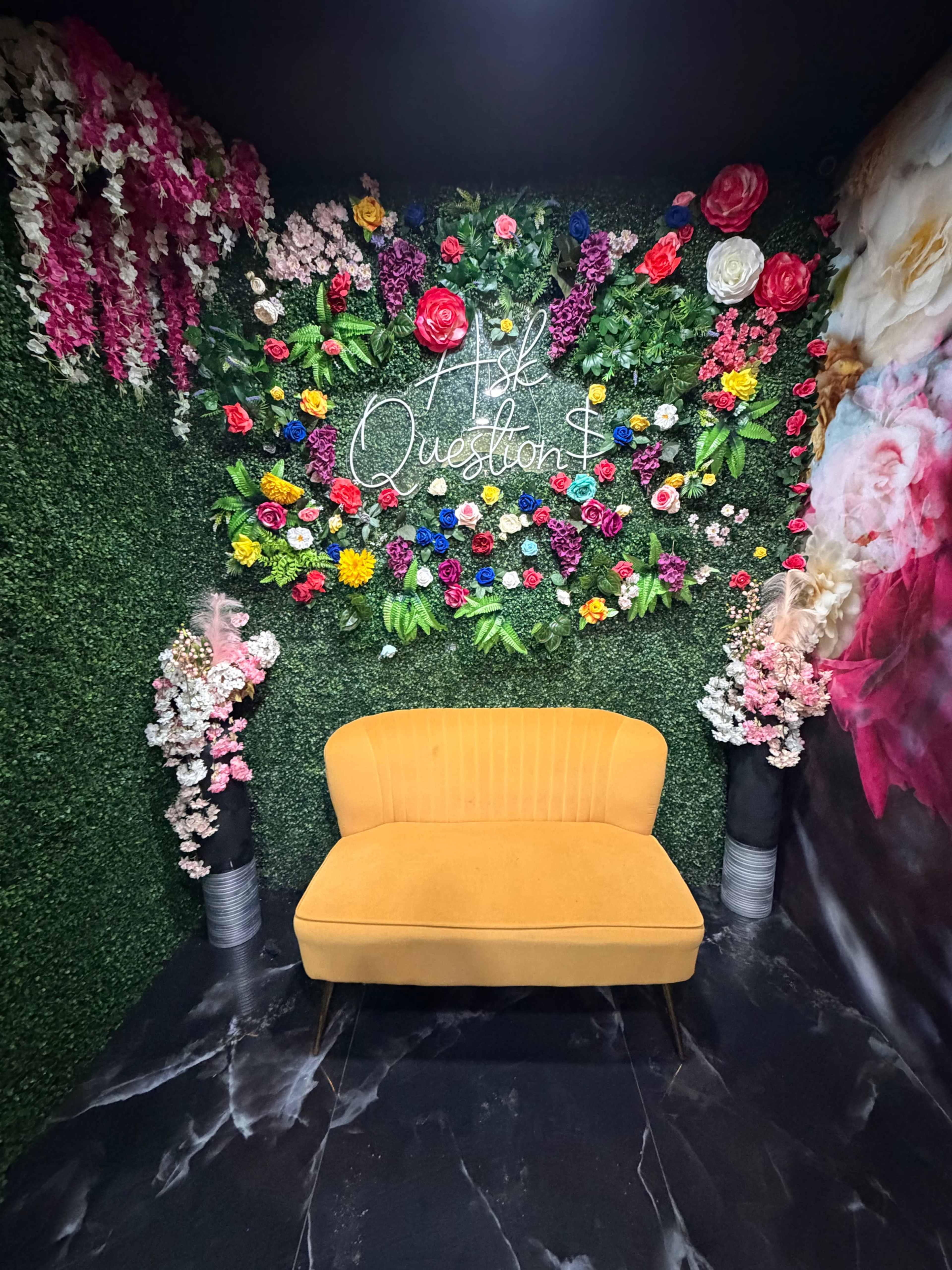 The image shows a cozy area featuring a yellow sofa in front of a vibrant floral backdrop with greenery and colorful artificial flowers.