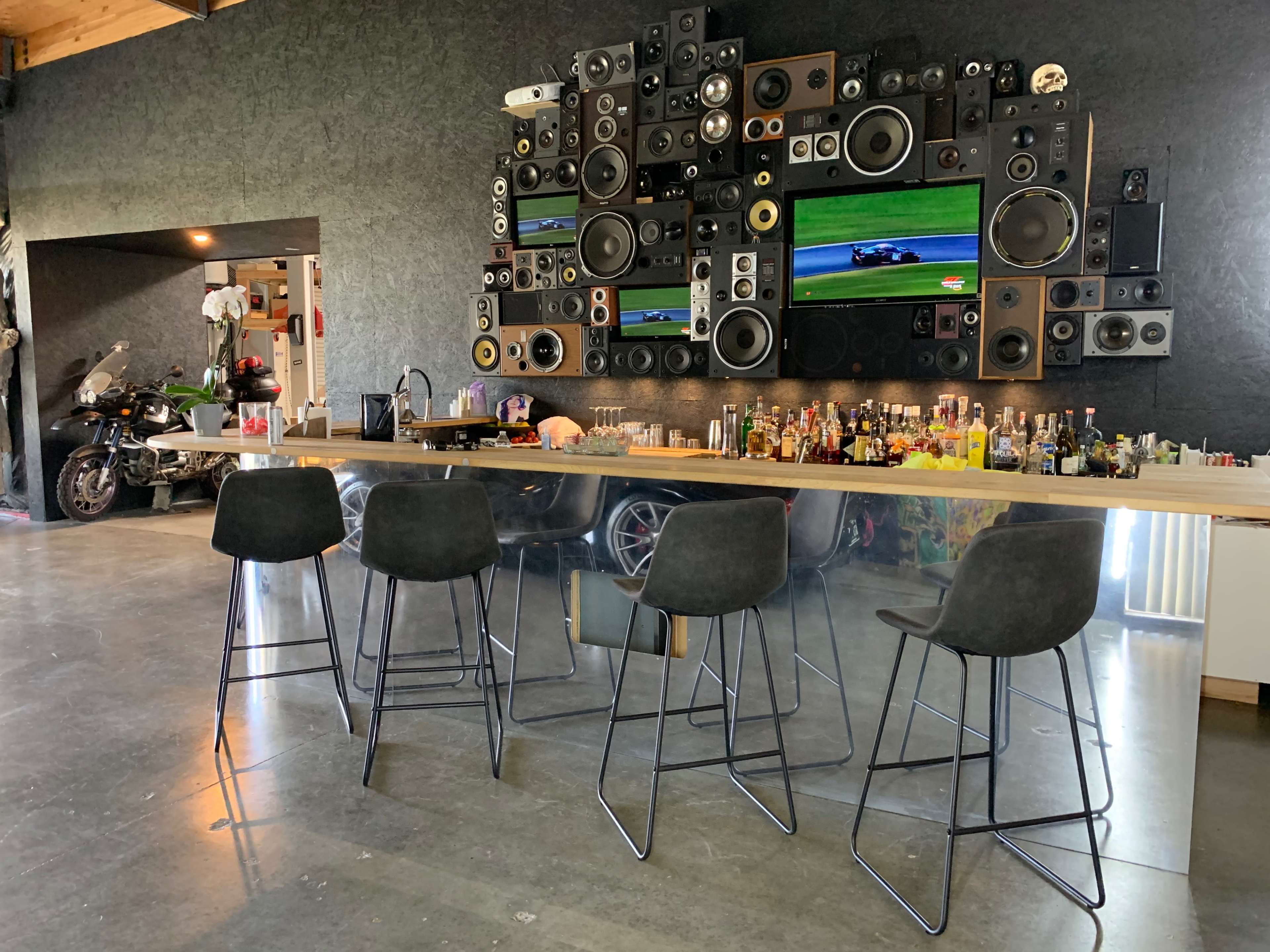 A modern bar area features a long counter with four stools, surrounded by an array of speakers and two televisions mounted on the wall, alongside a collection of liquor bottles.