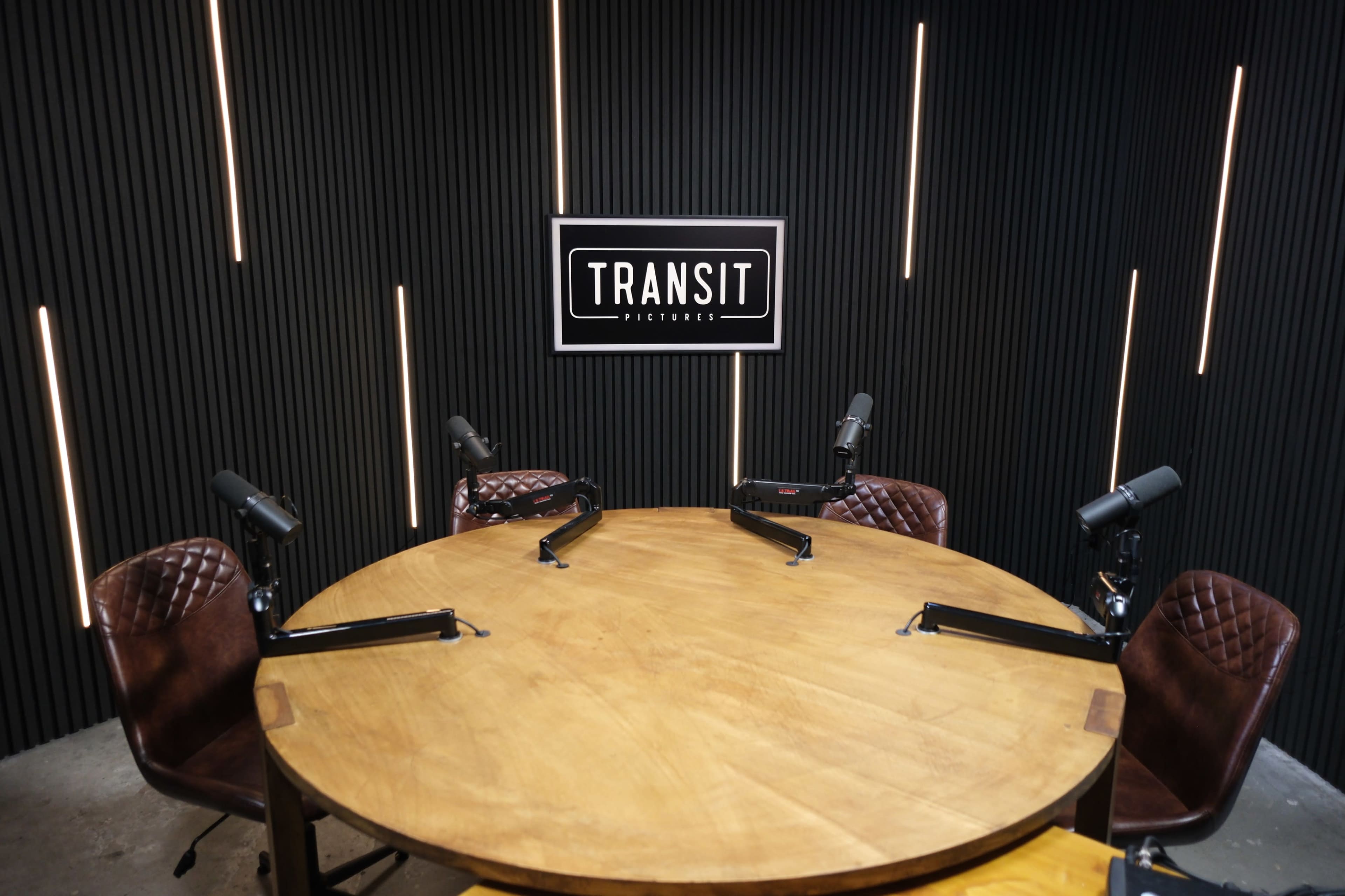 The image shows a recording studio with a circular wooden table surrounded by four chairs, each equipped with a microphone, and a large sign reading "TRANSIT PICTURES" on the wall.