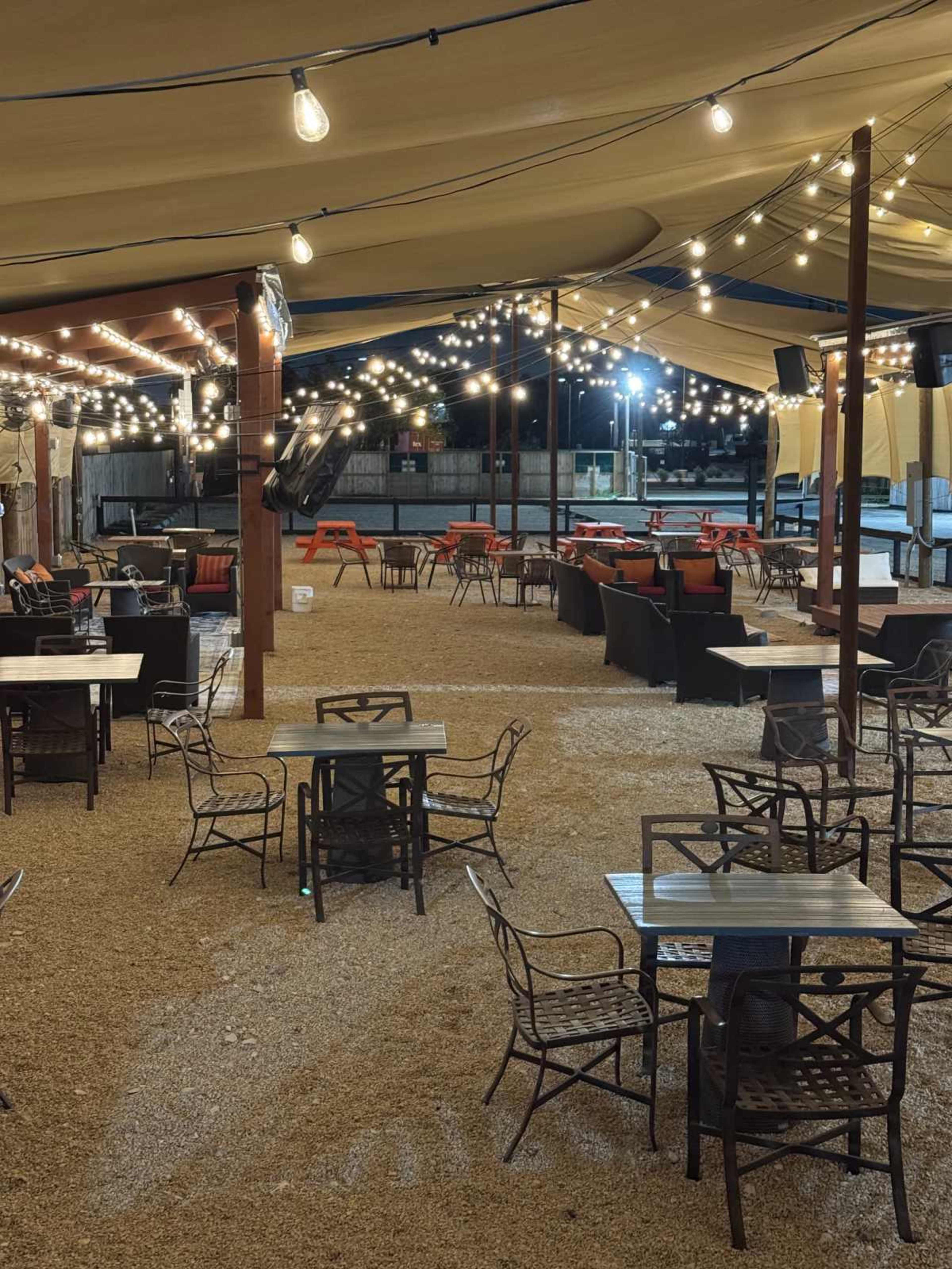 An outdoor dining area is illuminated by string lights, featuring several tables and seating arrangements under a large tent.