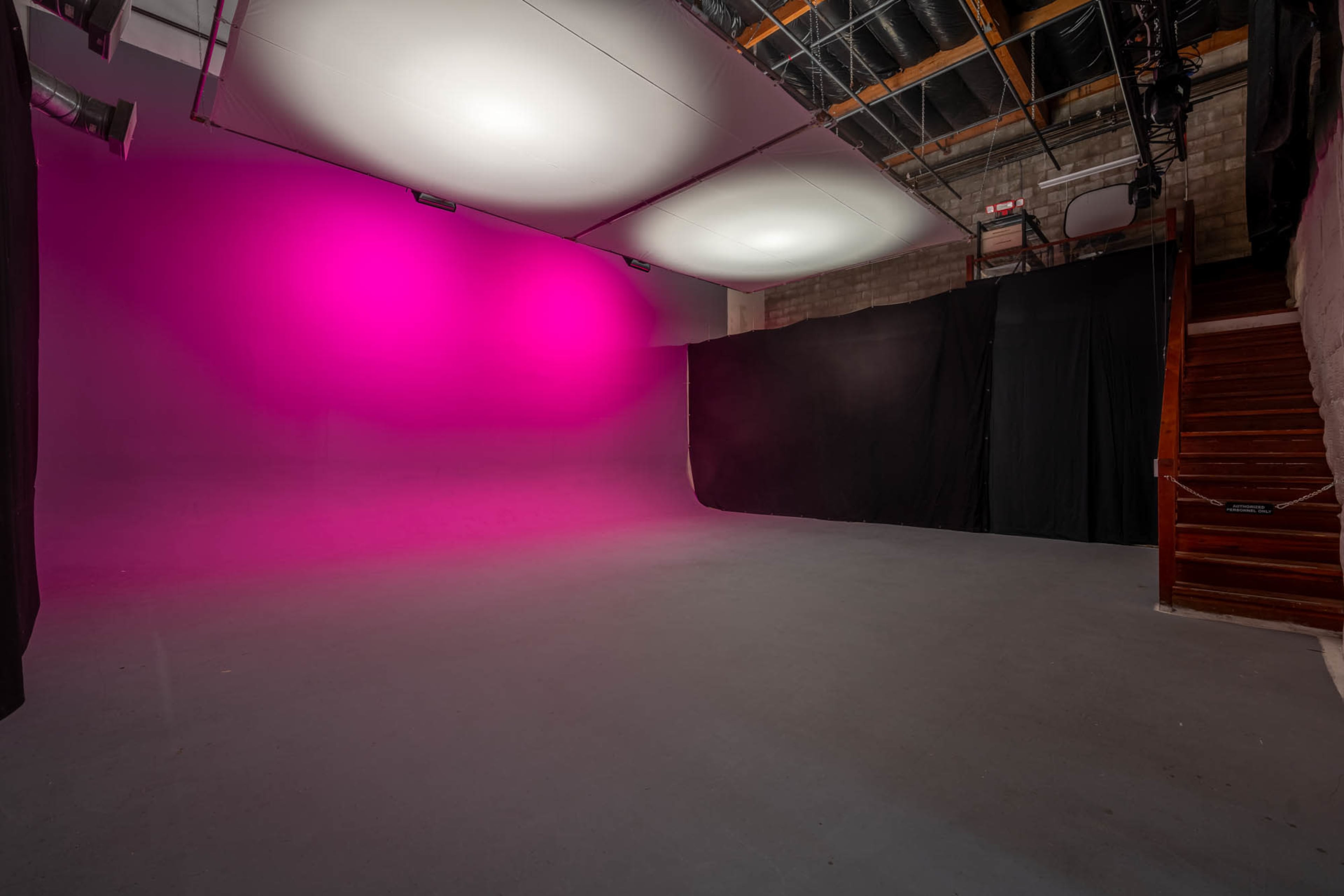 A spacious studio with a pink backdrop, black curtains, and a staircase on one side.